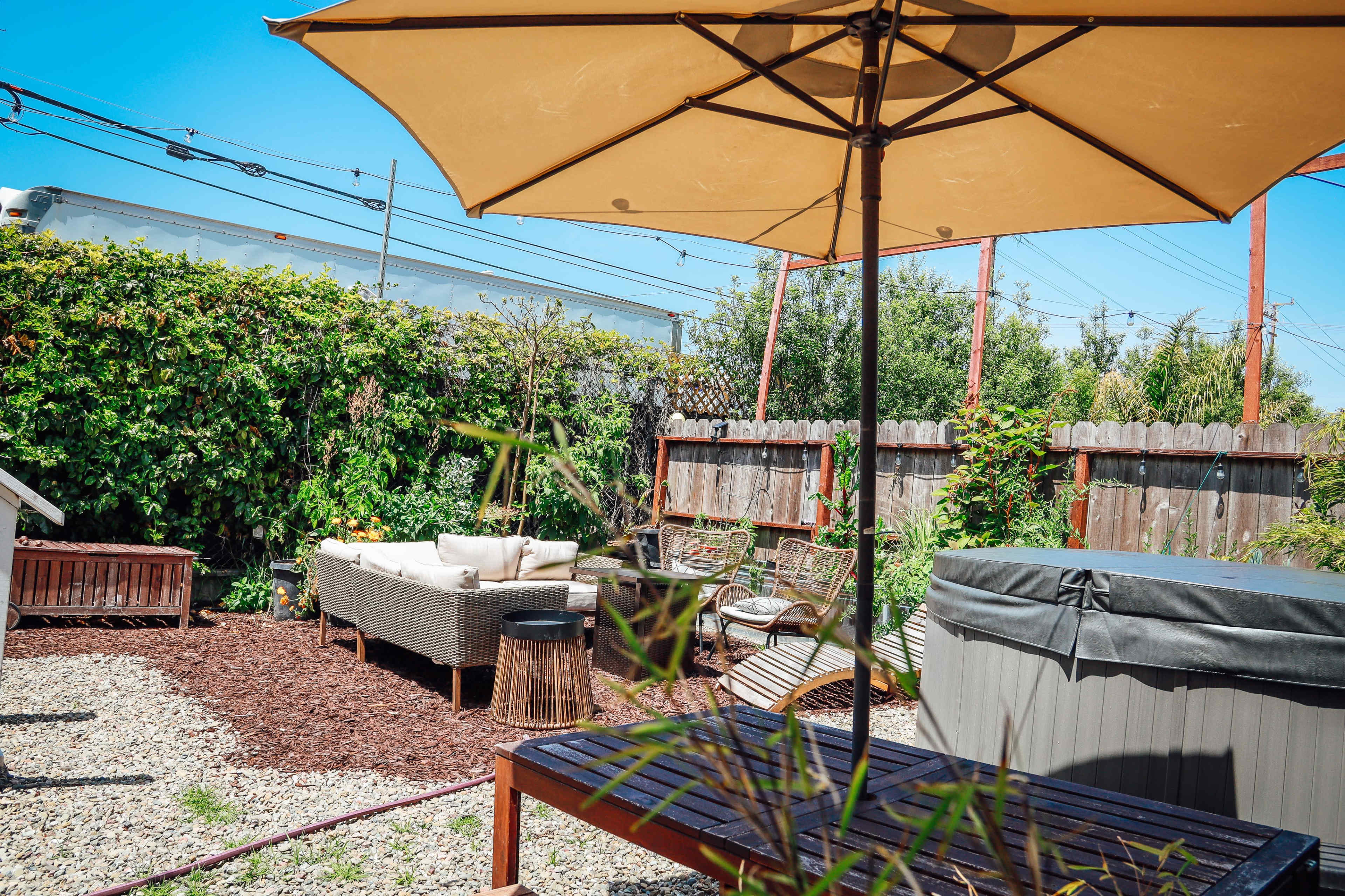 A backyard patio features a seating area with a couch and chairs, surrounded by greenery and a hot tub under a large umbrella.