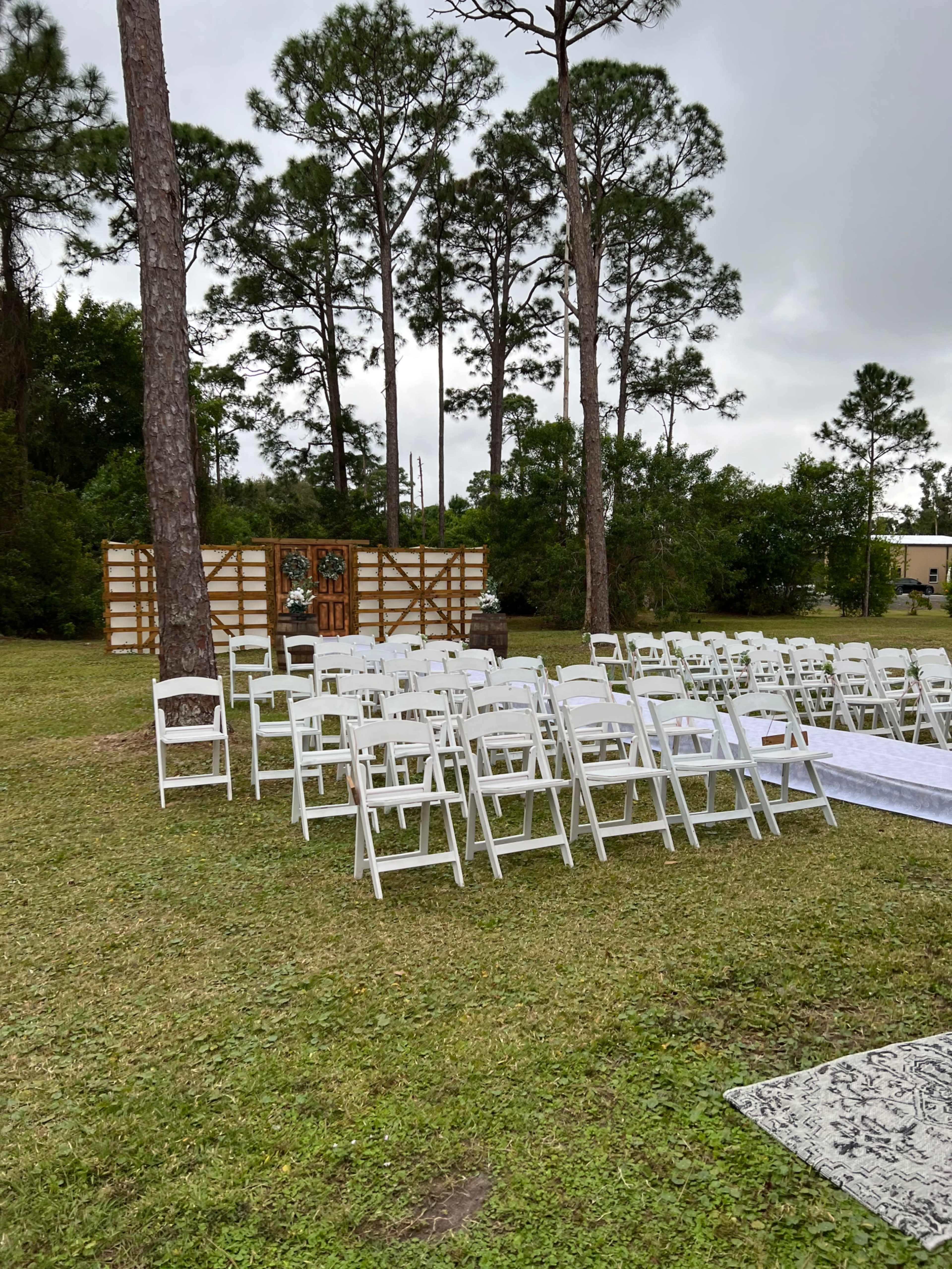 A row of white chairs is set up for a gathering in an outdoor area surrounded by trees, with a wooden backdrop visible in the background.