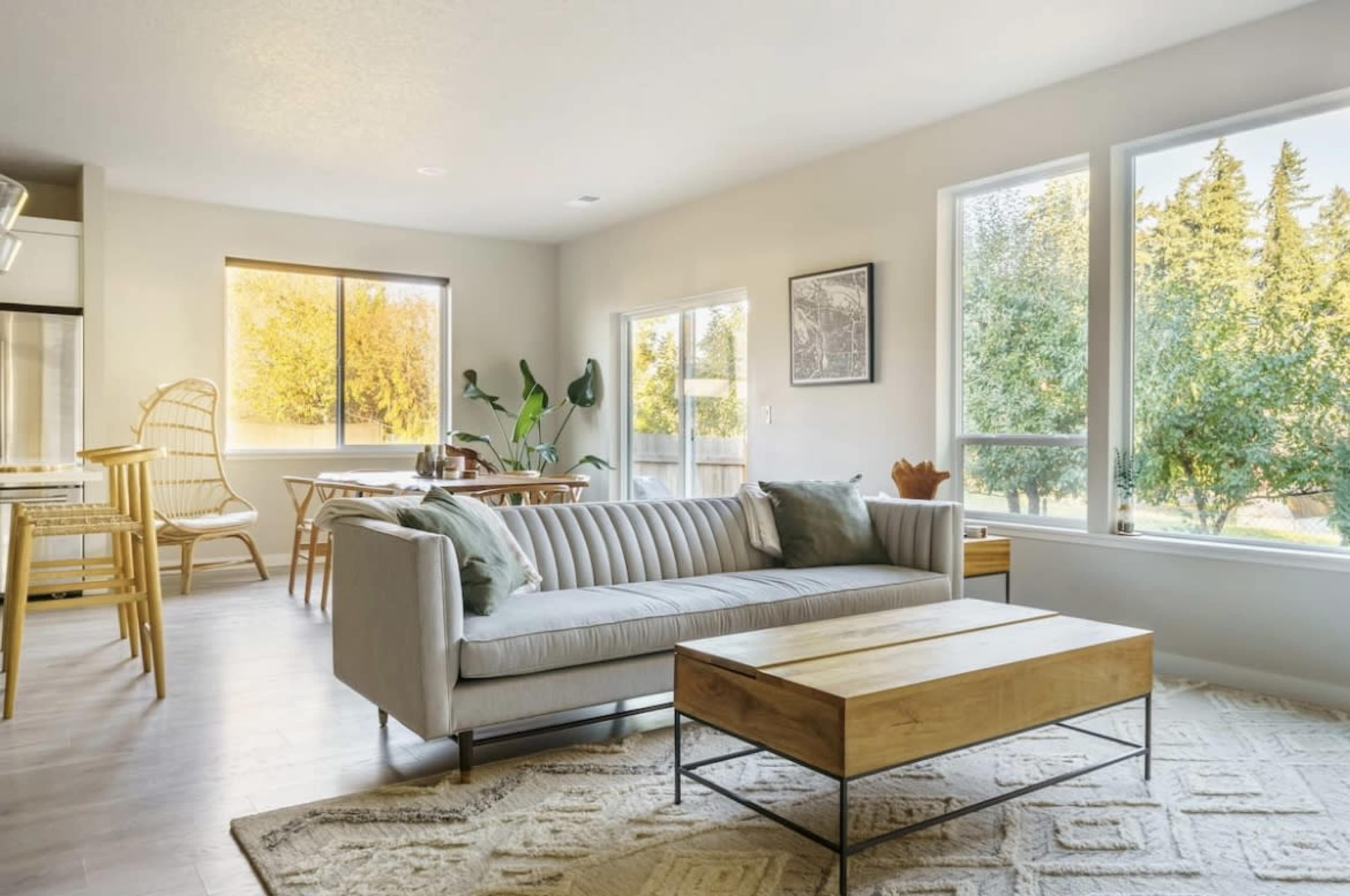 A modern living area features a striped sofa, a wooden coffee table, large windows, and a dining space connected to a kitchen.