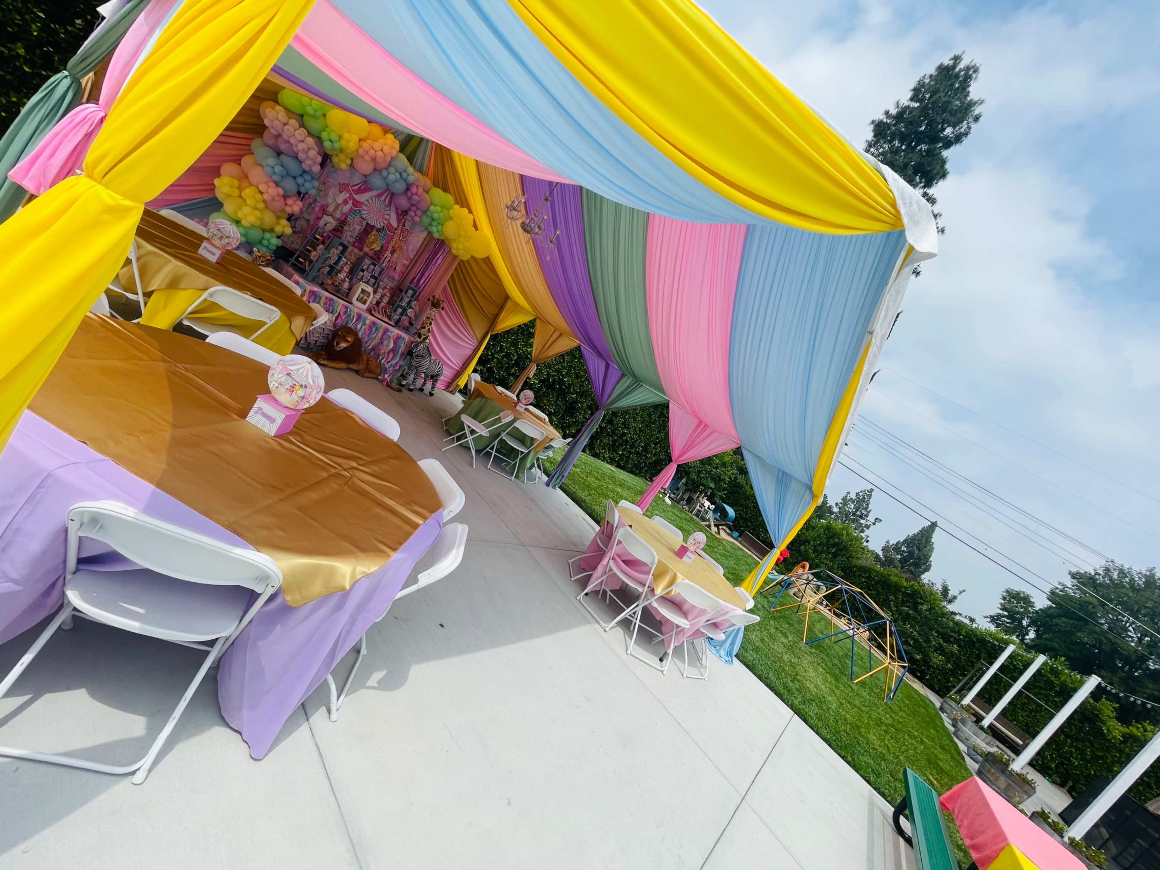 A colorful party tent is set up with decorated tables and a balloon backdrop in an outdoor setting.