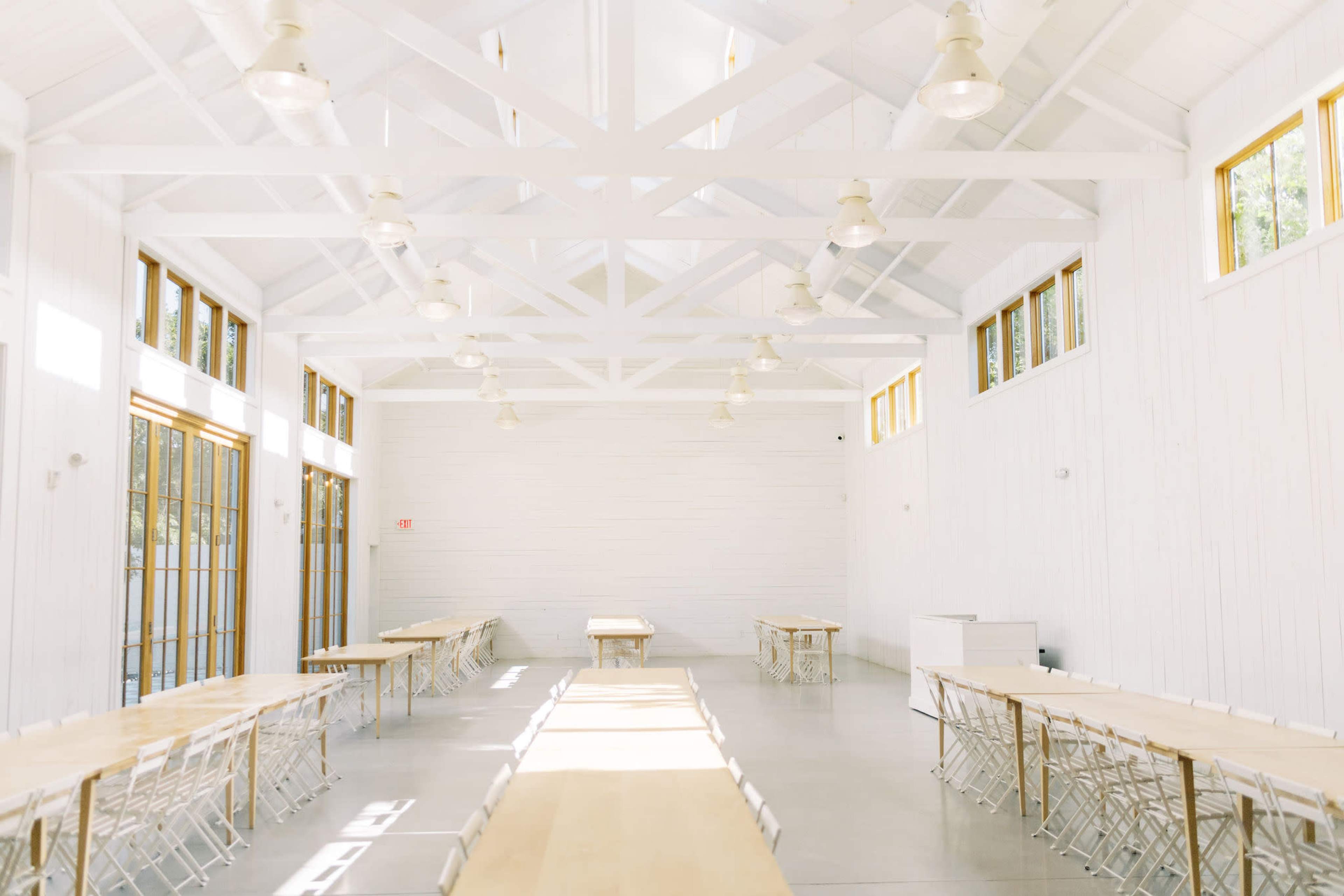 The image shows a bright, spacious interior of a hall with white walls, large windows, and long wooden tables arranged parallel to a central aisle.
