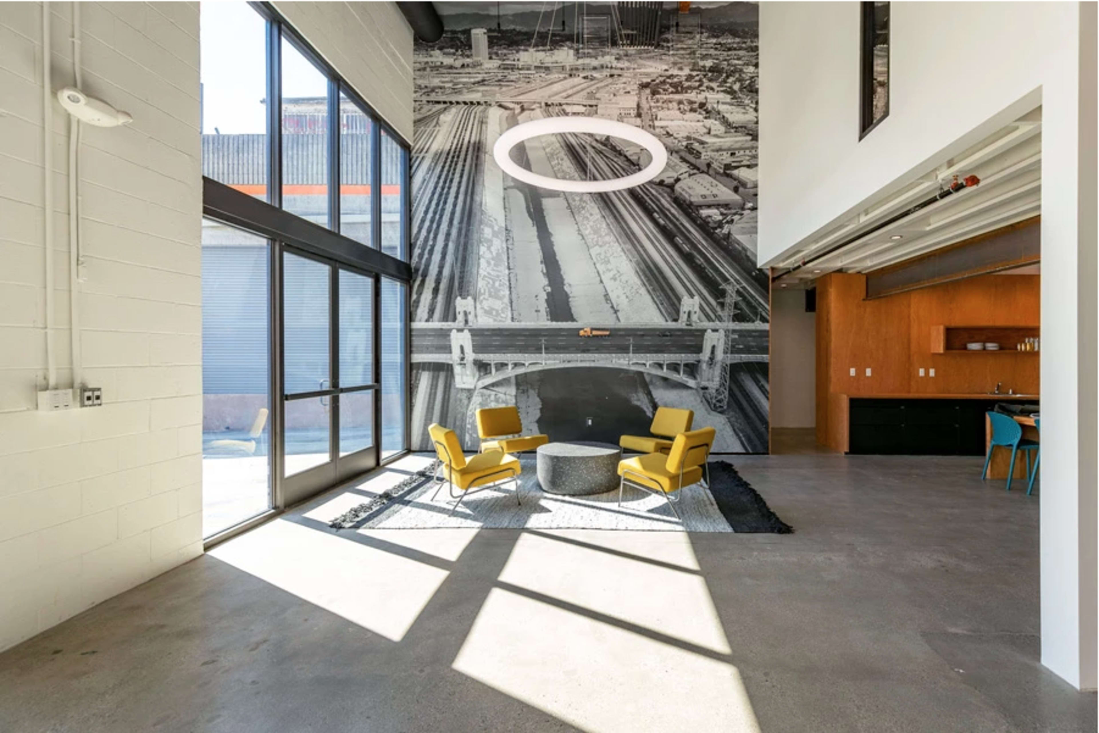 The image shows a modern interior with large windows, featuring two yellow chairs and a round coffee table on a rug, against a wall displaying a large black-and-white mural of a cityscape.