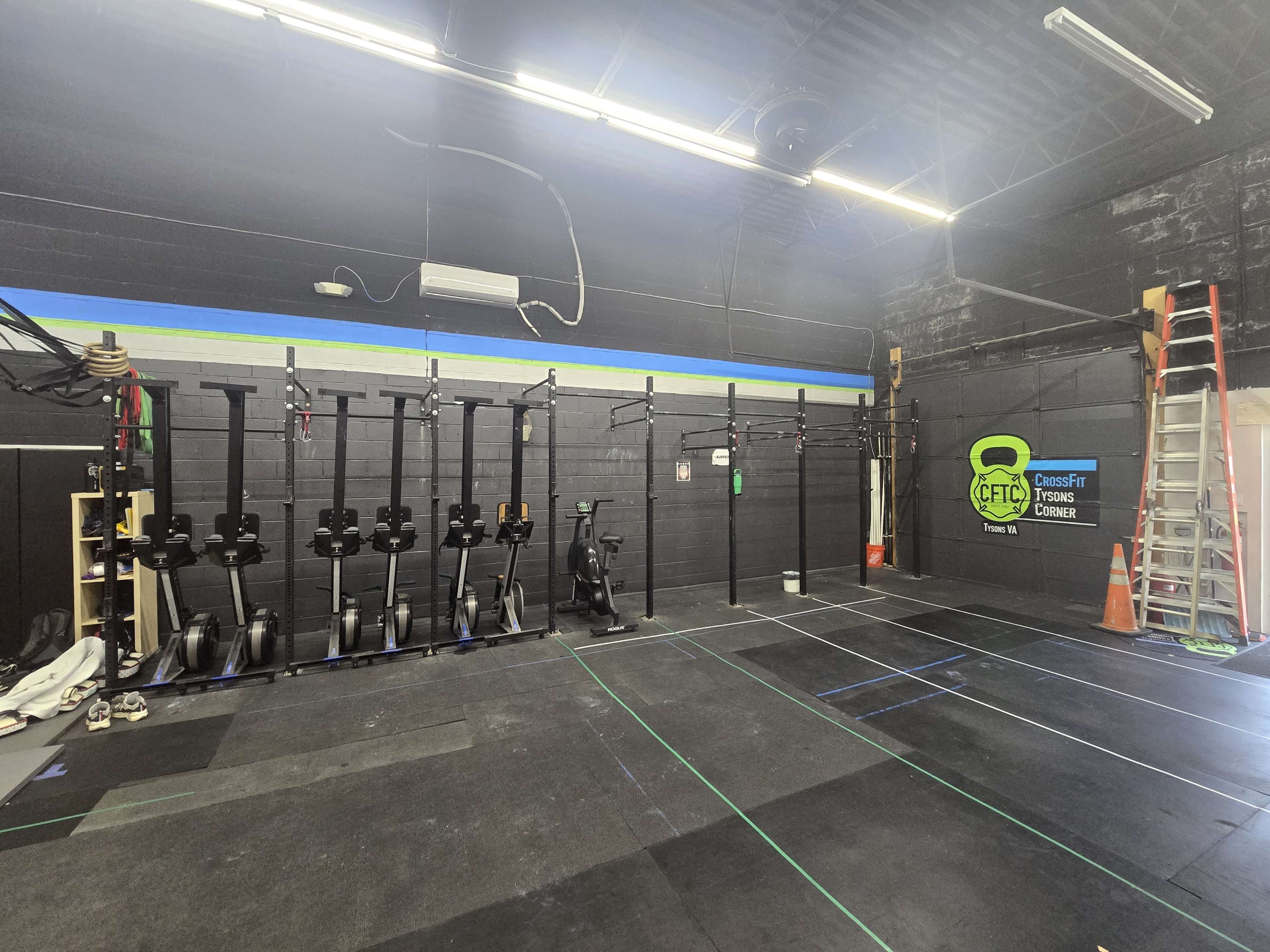 The image shows the interior of a gym with several rowing machines lined up against a black wall, along with fitness equipment and a ladder in the background.