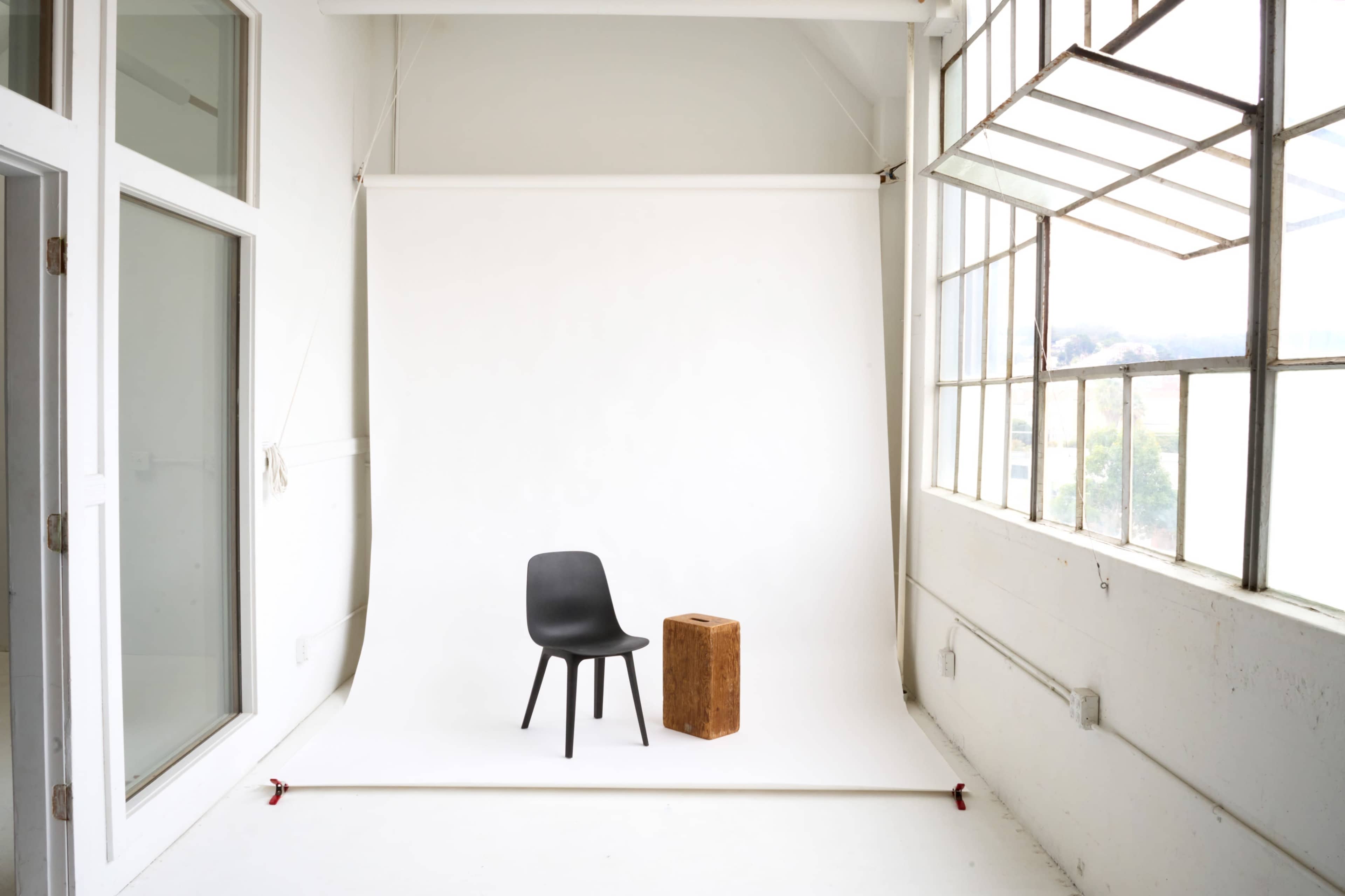 The image shows a minimalist studio space with a black chair and a wooden block set against a white backdrop and large windows.