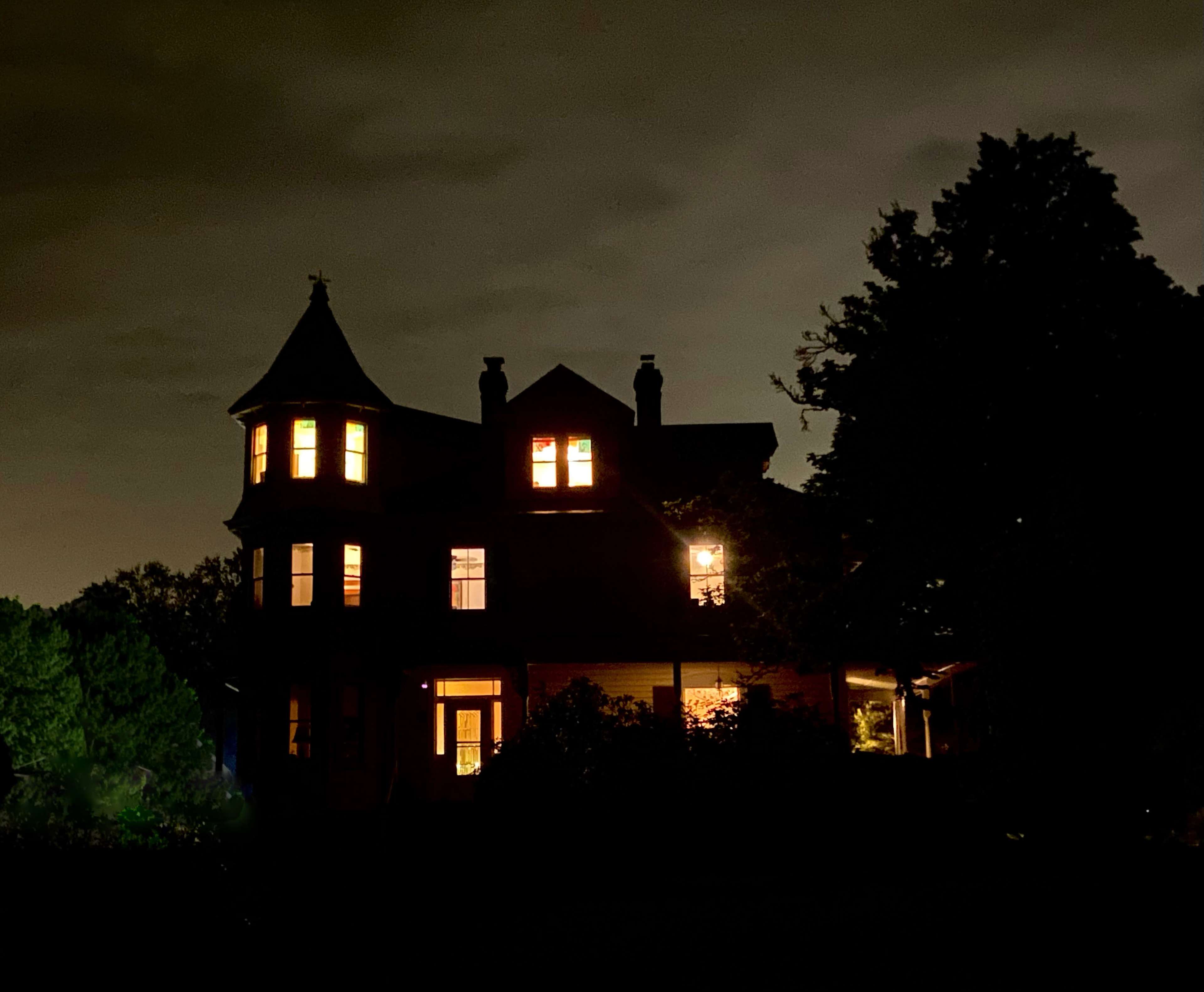 A Victorian-style house stands at night with illuminated windows under a dark sky.