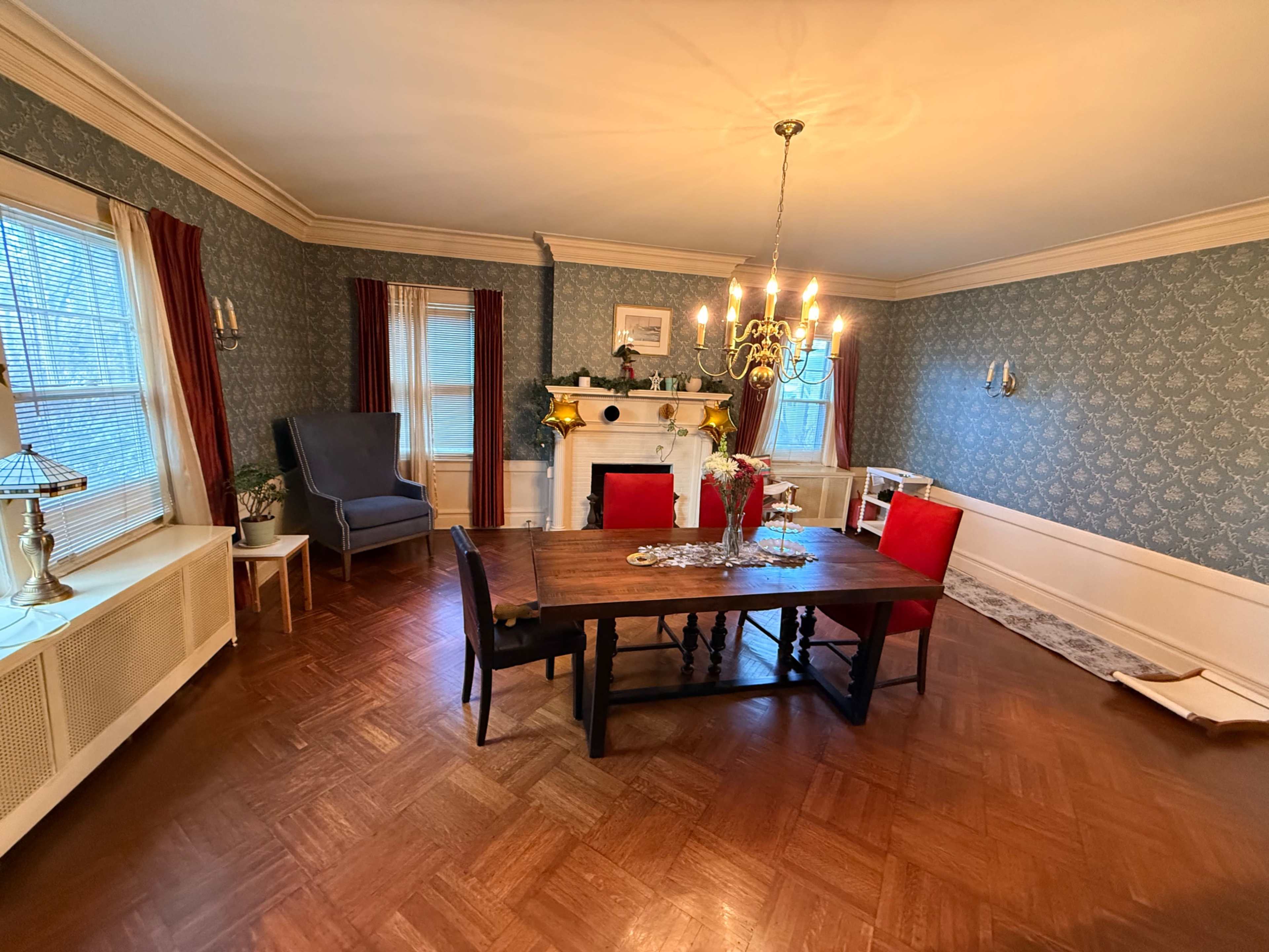 The image shows a spacious dining room with a large wooden table, a chandelier, and a fireplace, set against a patterned wallpaper.