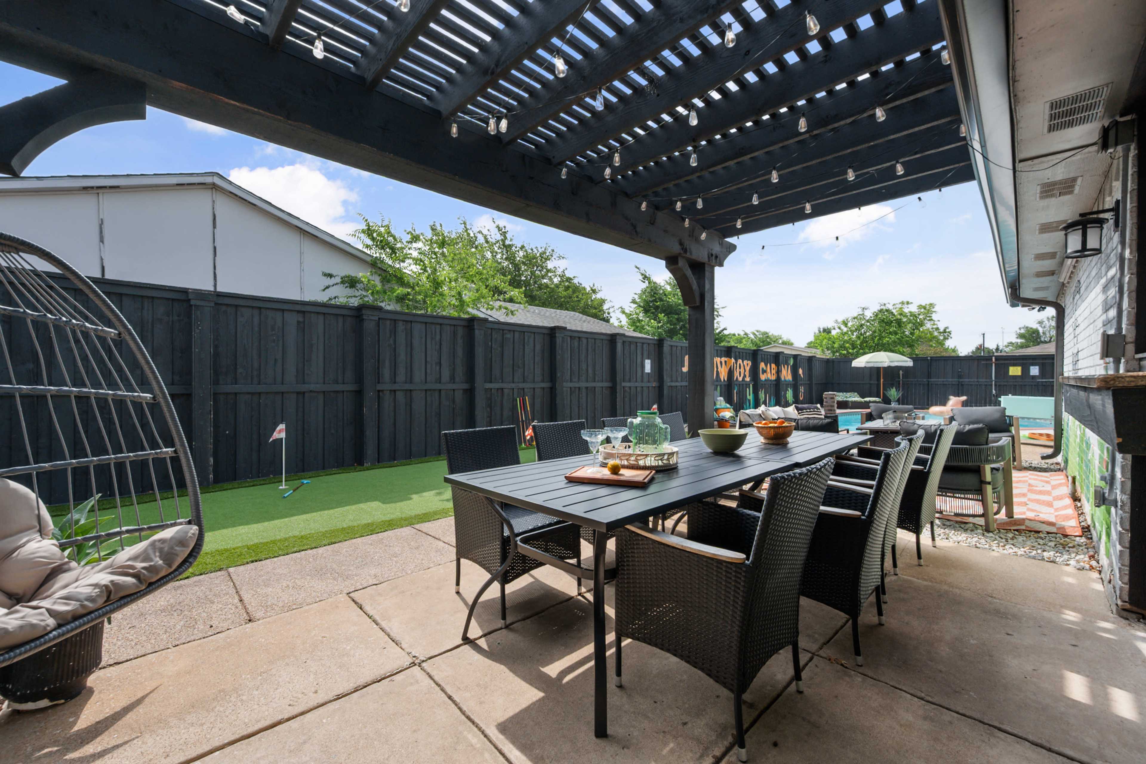 The image shows a backyard patio with a large dining table, surrounded by chairs, and a green lawn area behind a black wooden fence.