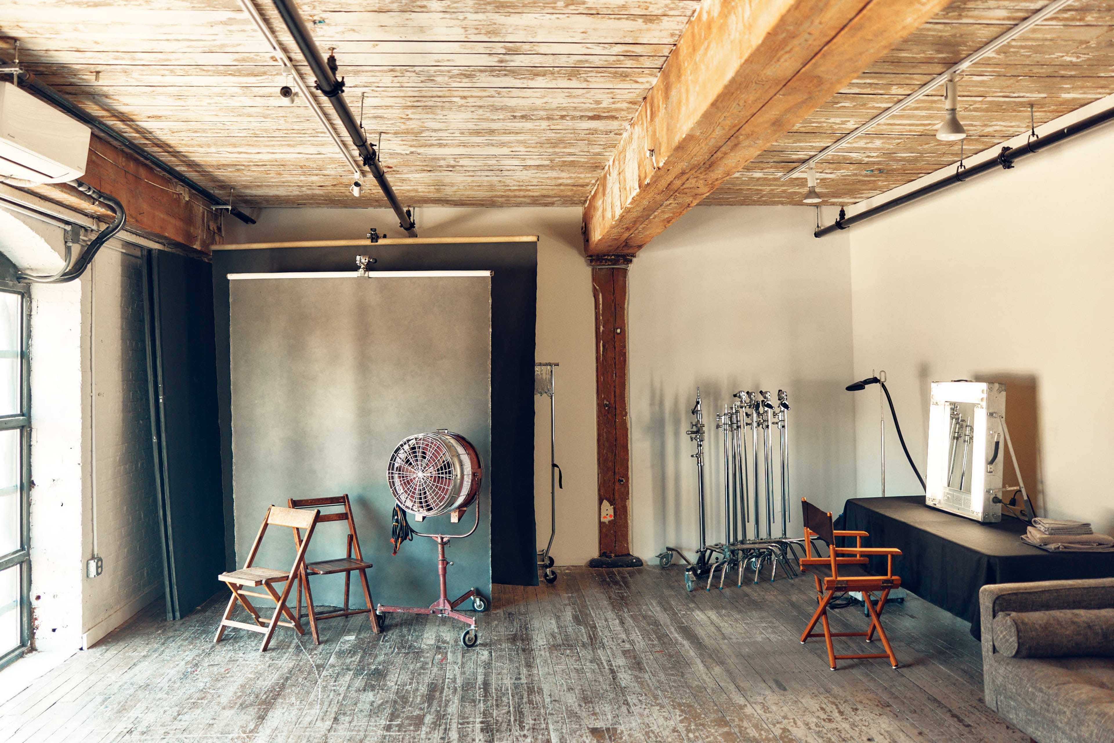 The image shows a spacious, well-lit studio with wooden beams, a gray backdrop, chairs, a fan, and photography equipment neatly arranged.