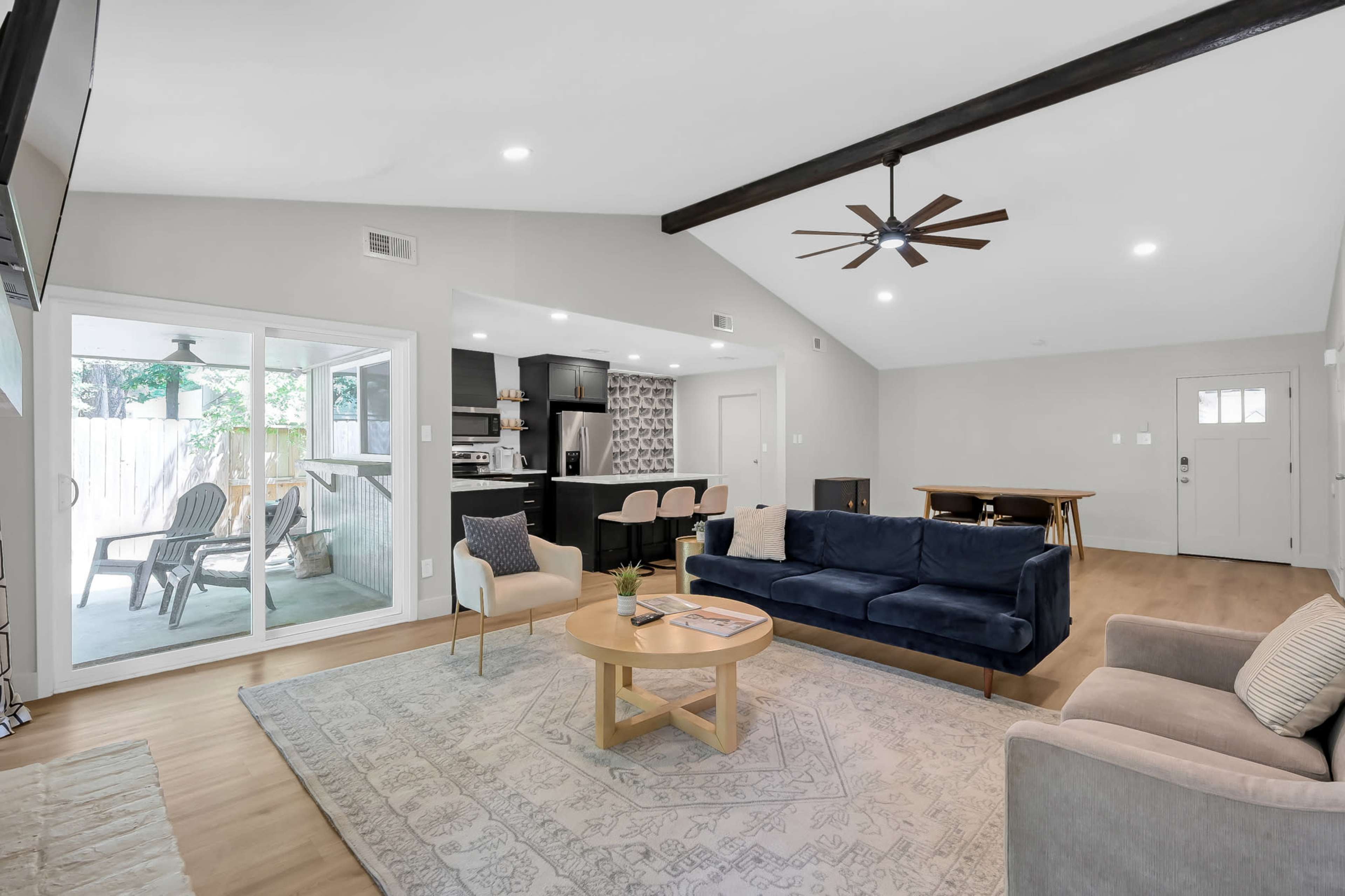 The living room features a navy blue sofa, a round coffee table, and a ceiling fan, with an open view into a kitchen area and a glass door leading to an outside porch.