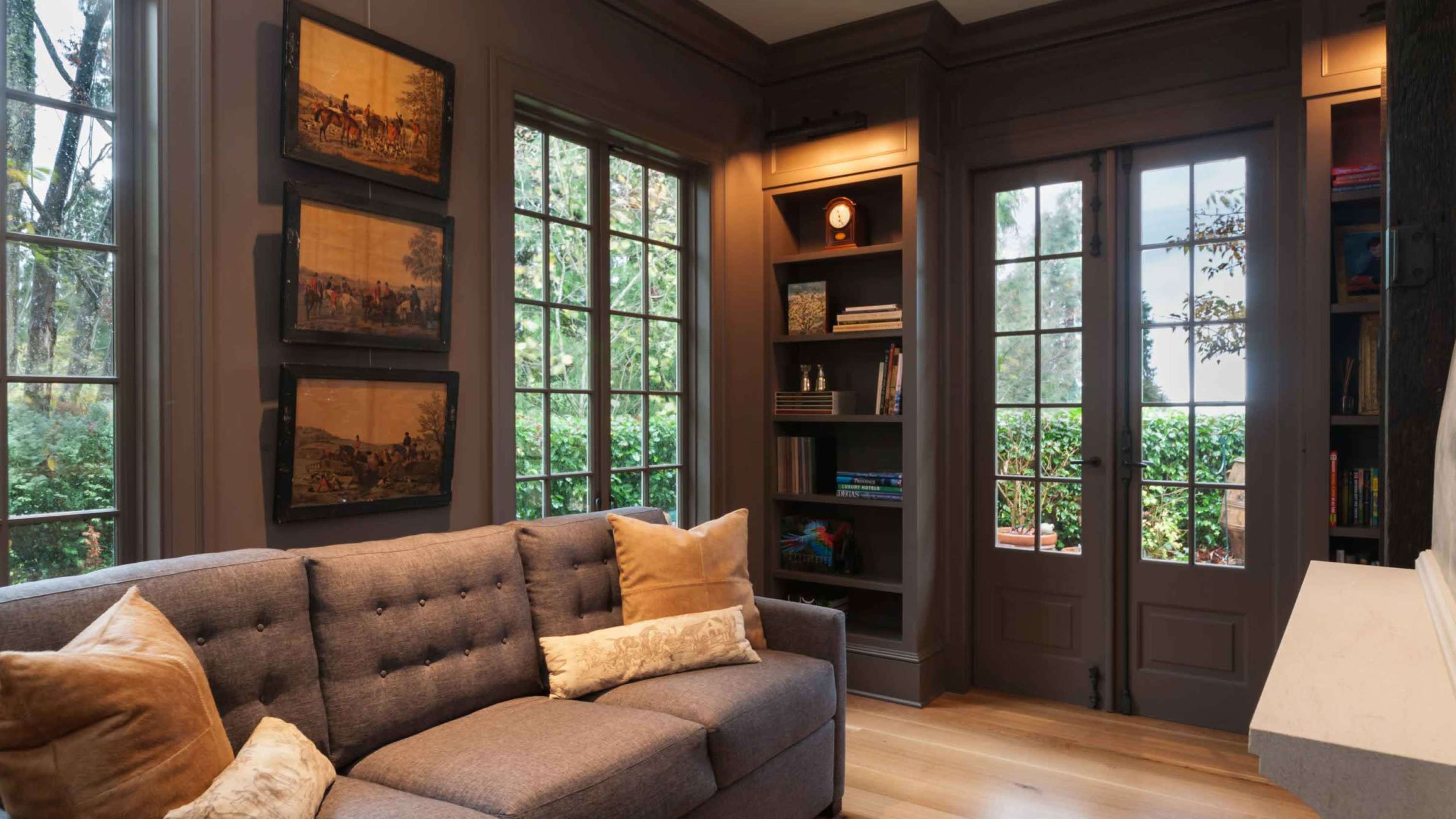 The image shows a cozy room featuring a gray sofa, wooden bookshelves, framed artwork on the walls, and large windows with a view of greenery outside.