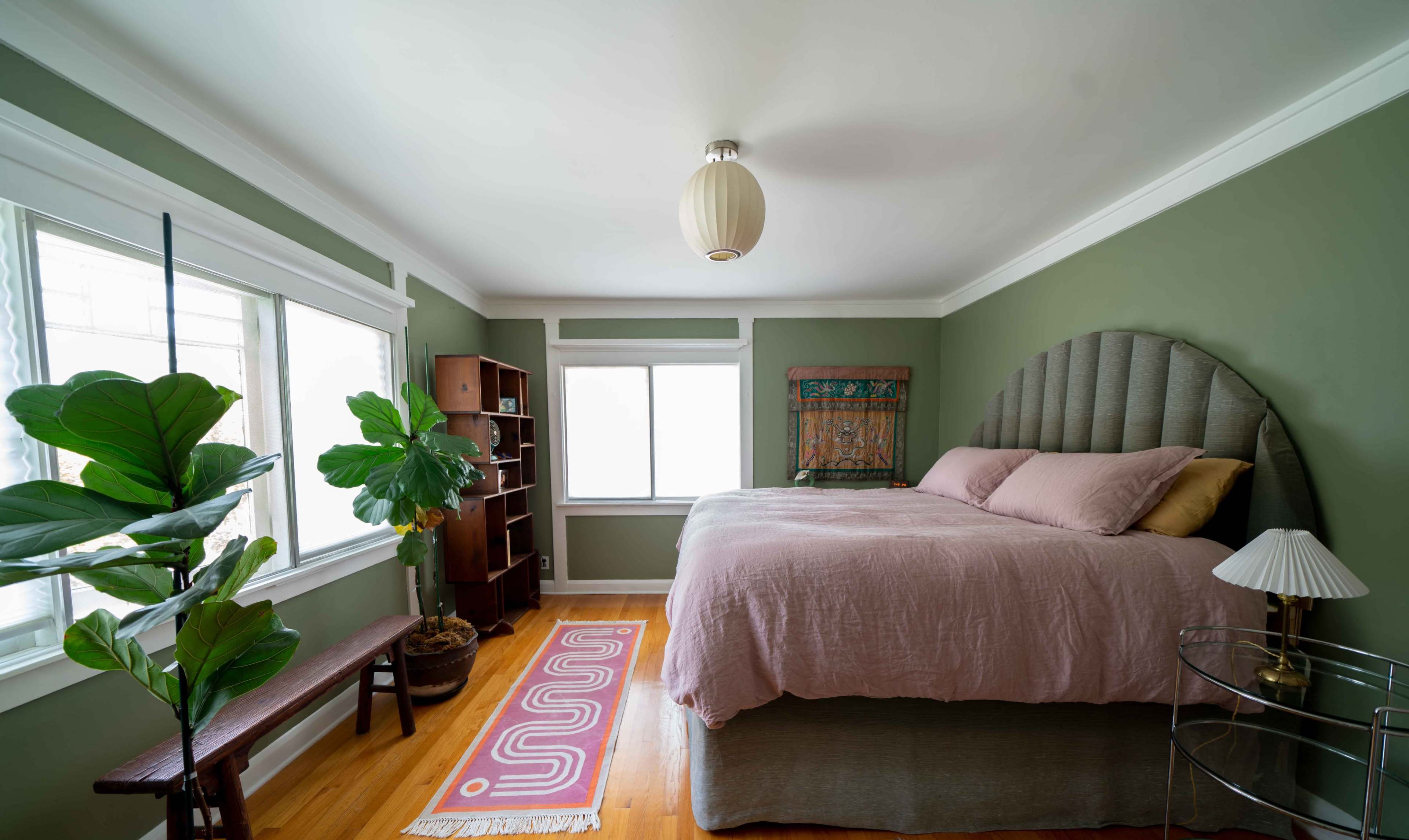 A neatly arranged bedroom features a large bed with a pink coverlet, a green accent wall, a wooden bench, and a potted plant near the windows.