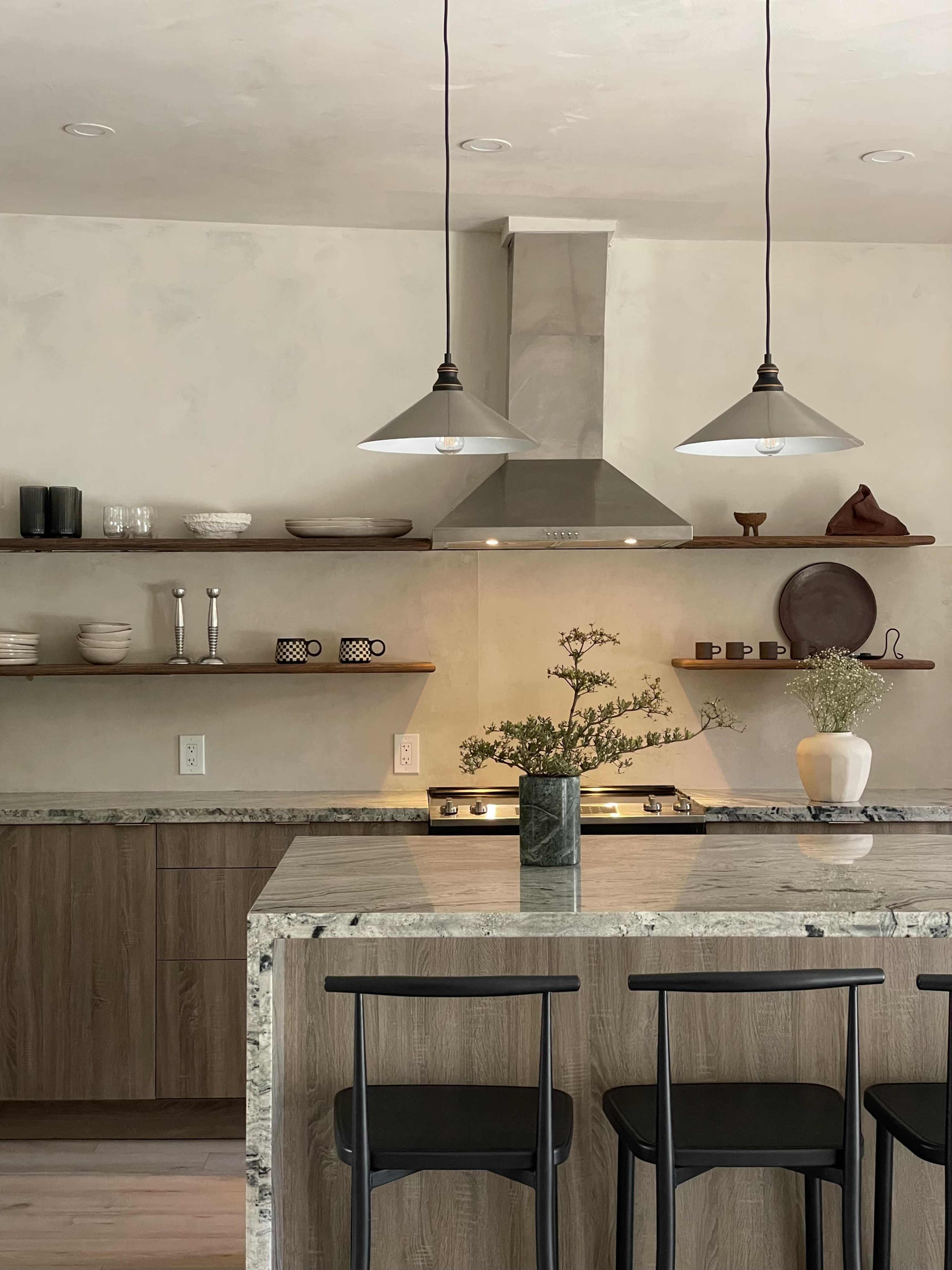 A modern kitchen features a central island with bar stools, pendant lighting, and open shelving displaying various dishes and decor.