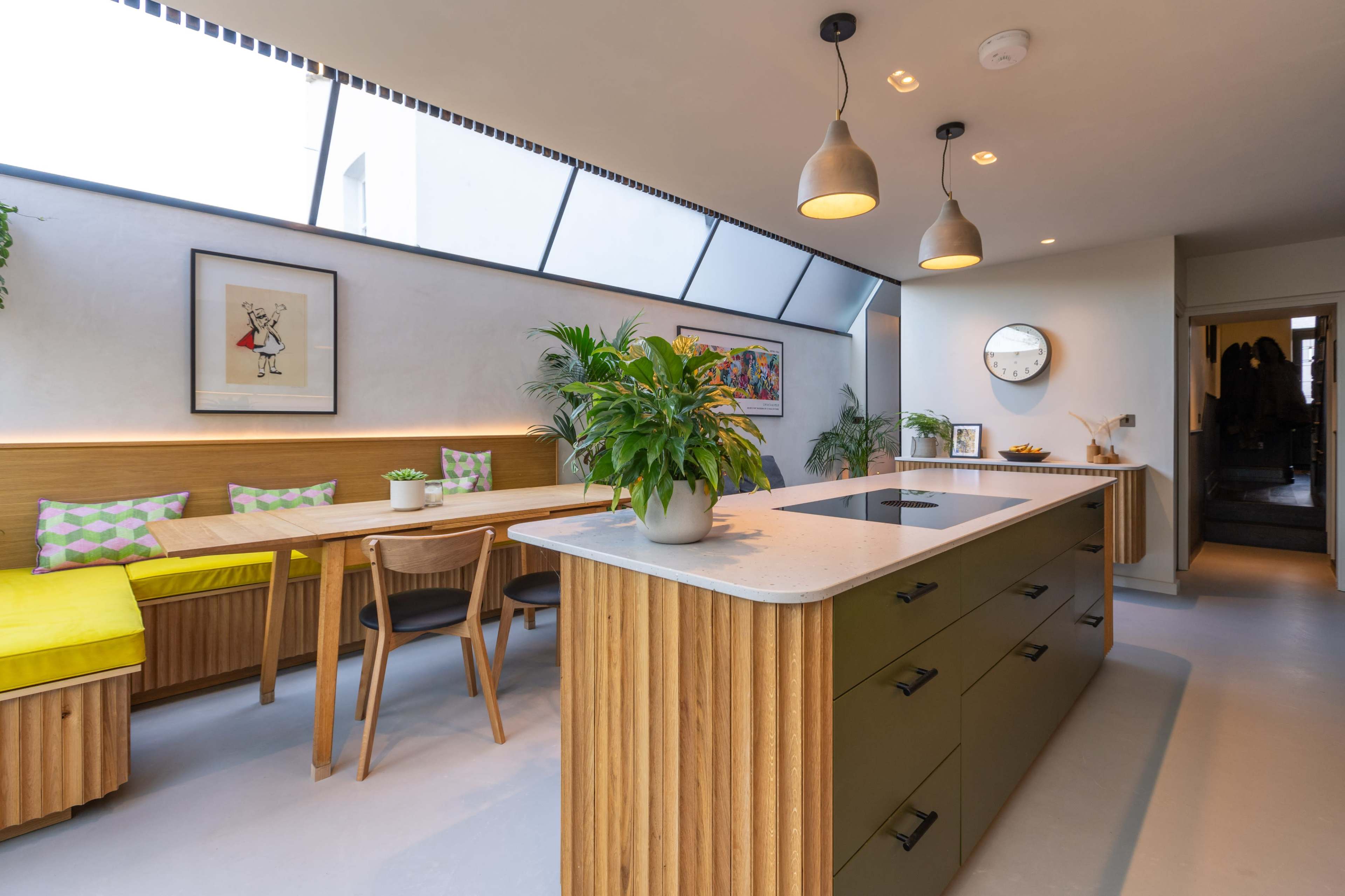 The image shows a modern kitchen with an island, a wooden dining table, and a built-in seating area illuminated by natural light from a large window.