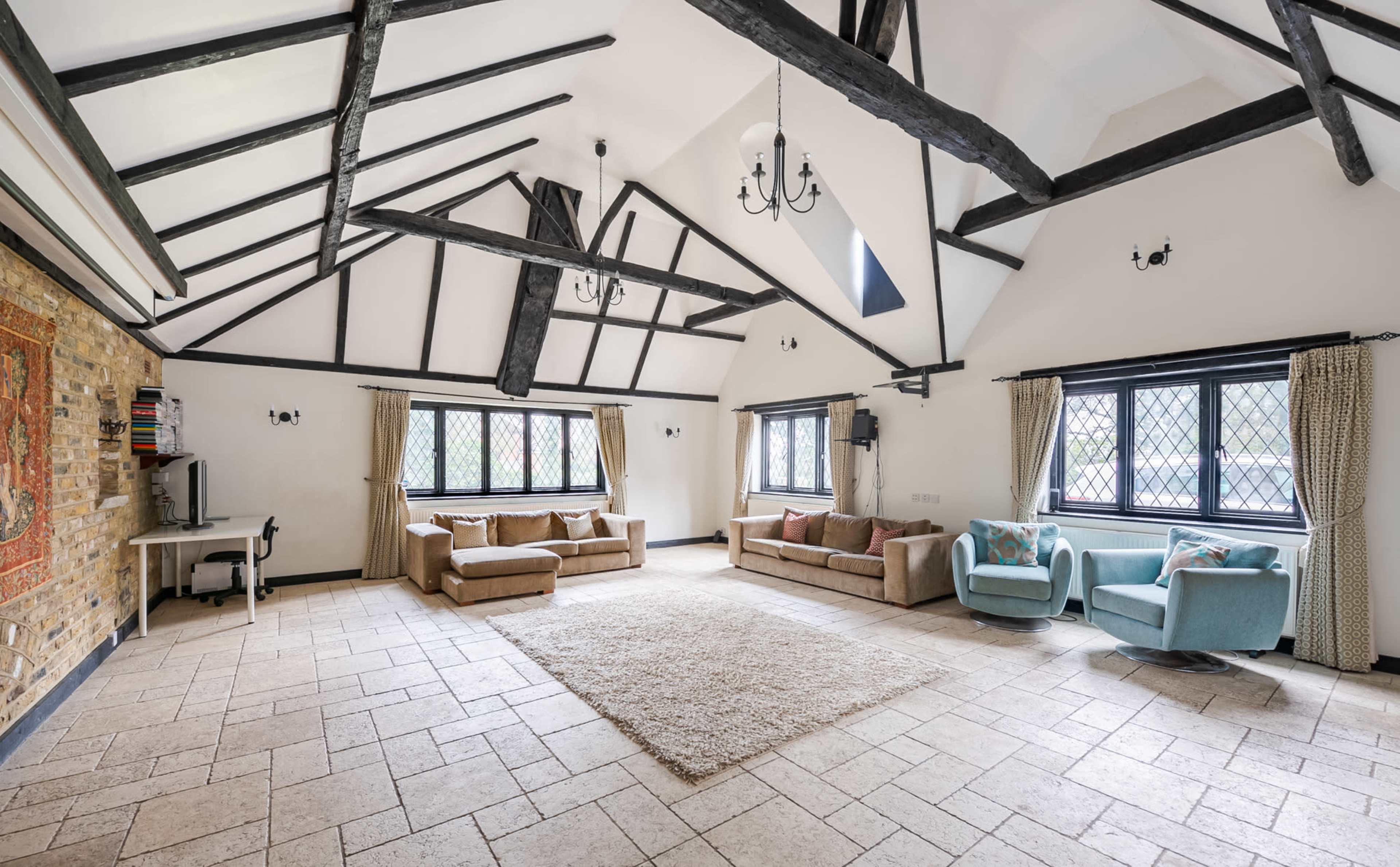 The image shows a spacious living area with high ceilings, exposed beams, large windows, and a mix of seating options, including sofas and armchairs, on a stone-tiled floor.