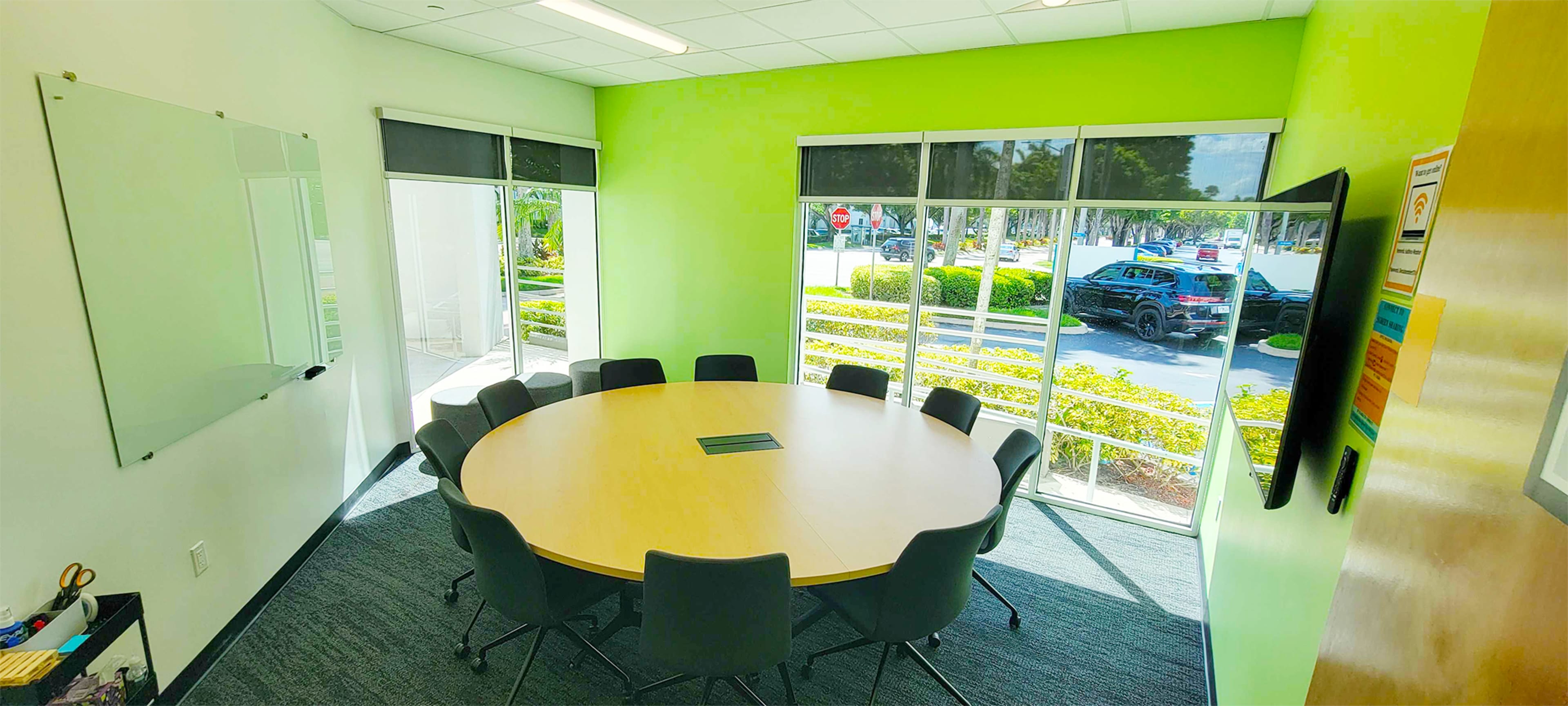 A conference room features a round table with chairs arranged around it, large windows overlooking a landscaped area, and a whiteboard on one wall.