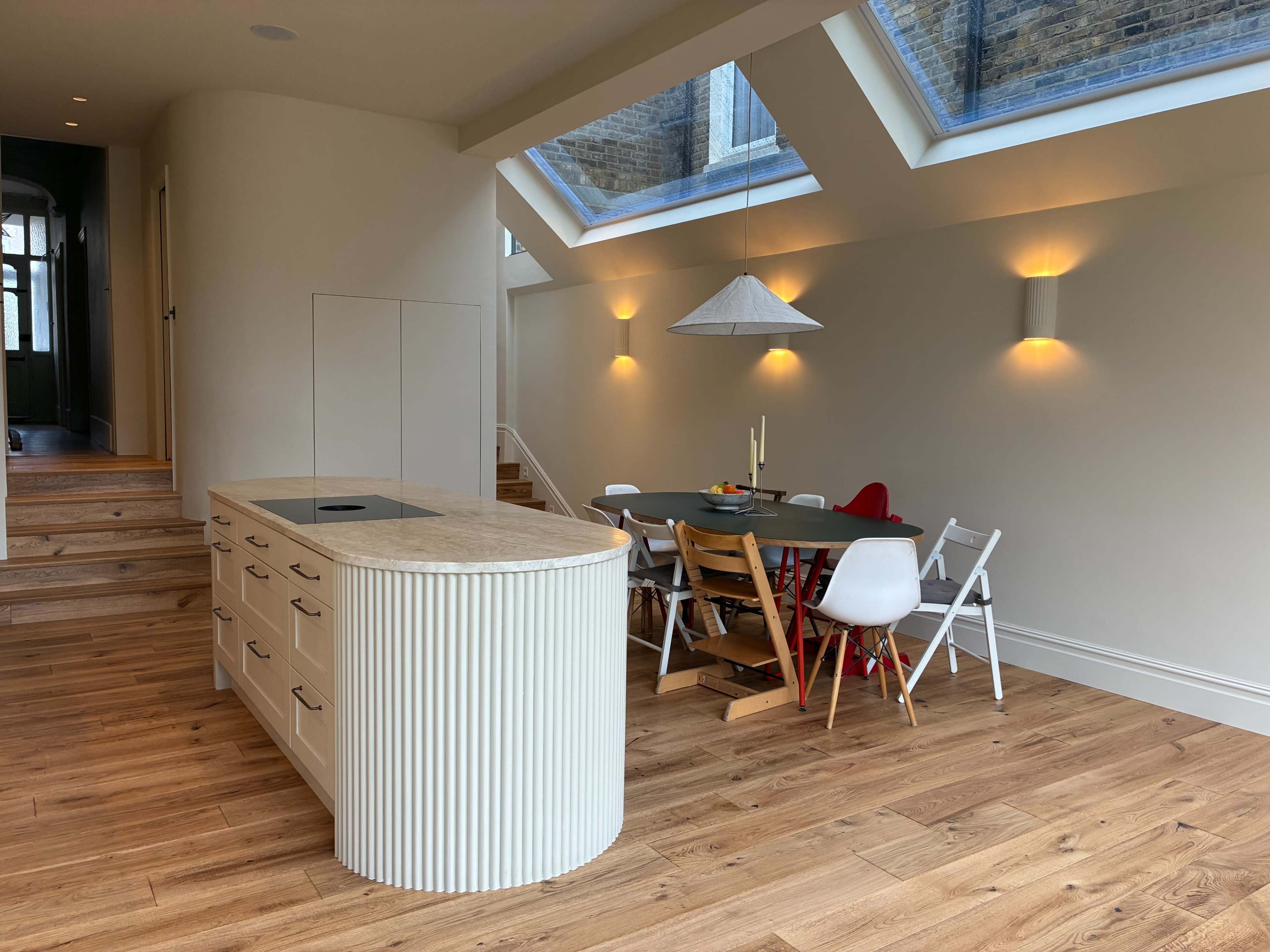 The image shows a modern kitchen with an island and a dining area featuring a table surrounded by various chairs, all under skylights in a bright, open space.