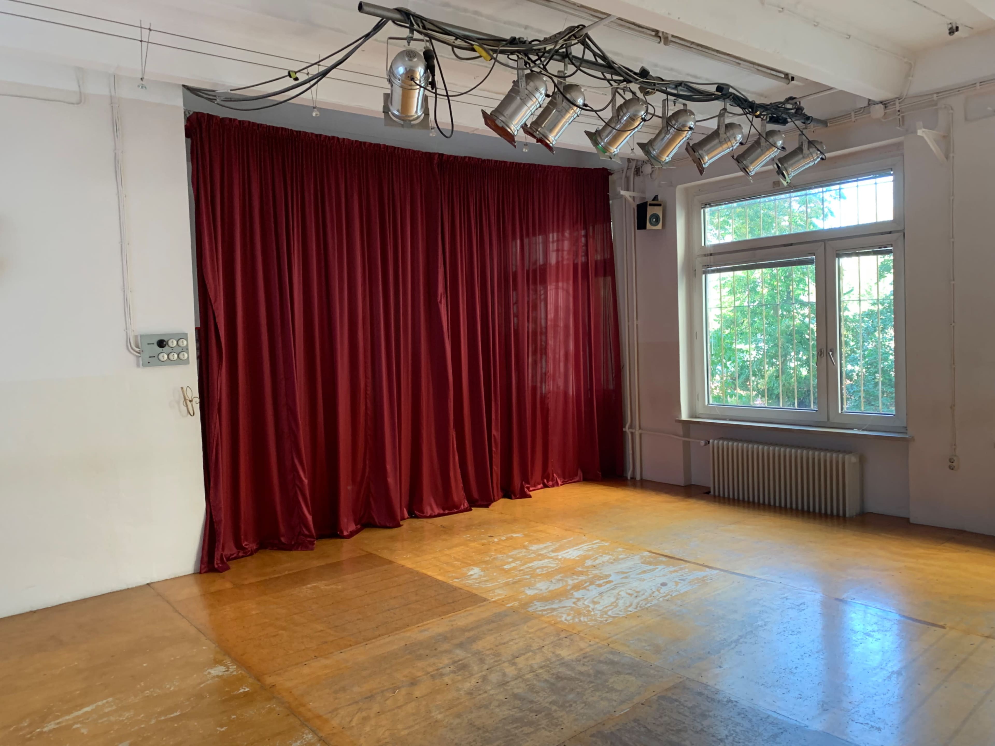 An empty room with a wooden floor, red curtains at one end, and overhead lighting fixtures.