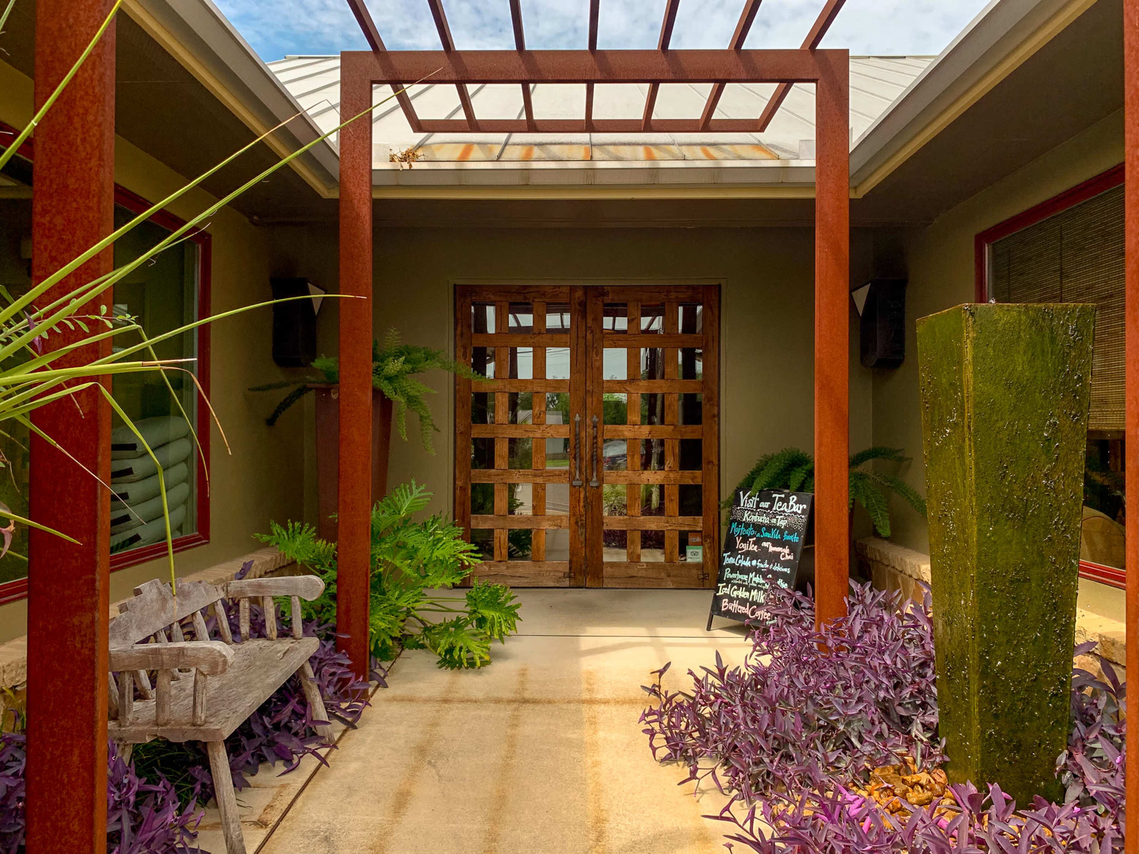 The entrance features a wooden door flanked by planters and a bench, beneath a pergola with a sign detailing services on the side.