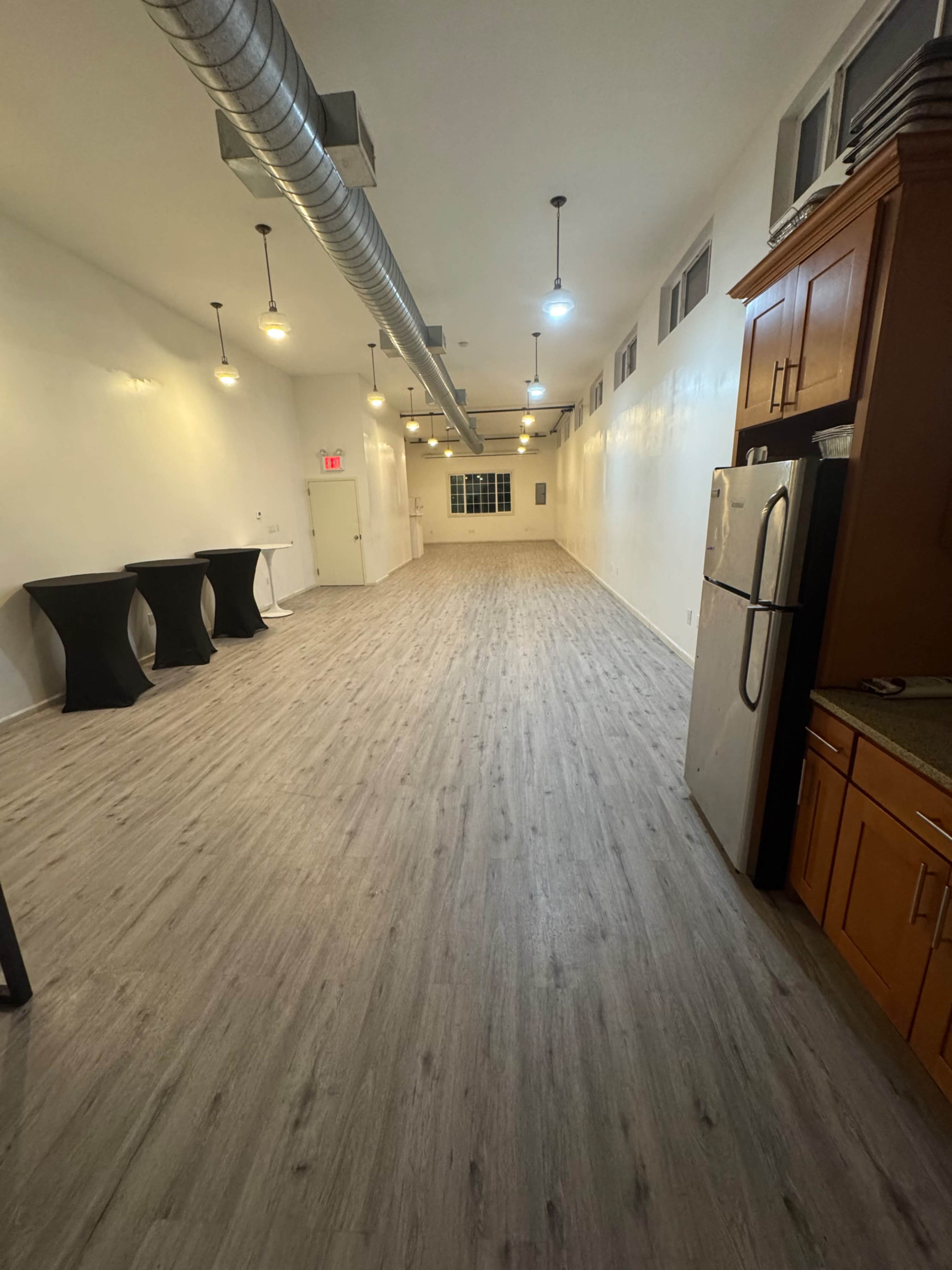 The image shows a long, empty room with light-colored wooden flooring, exposed ductwork along the ceiling, and a kitchenette area with a refrigerator and cabinets on one side.