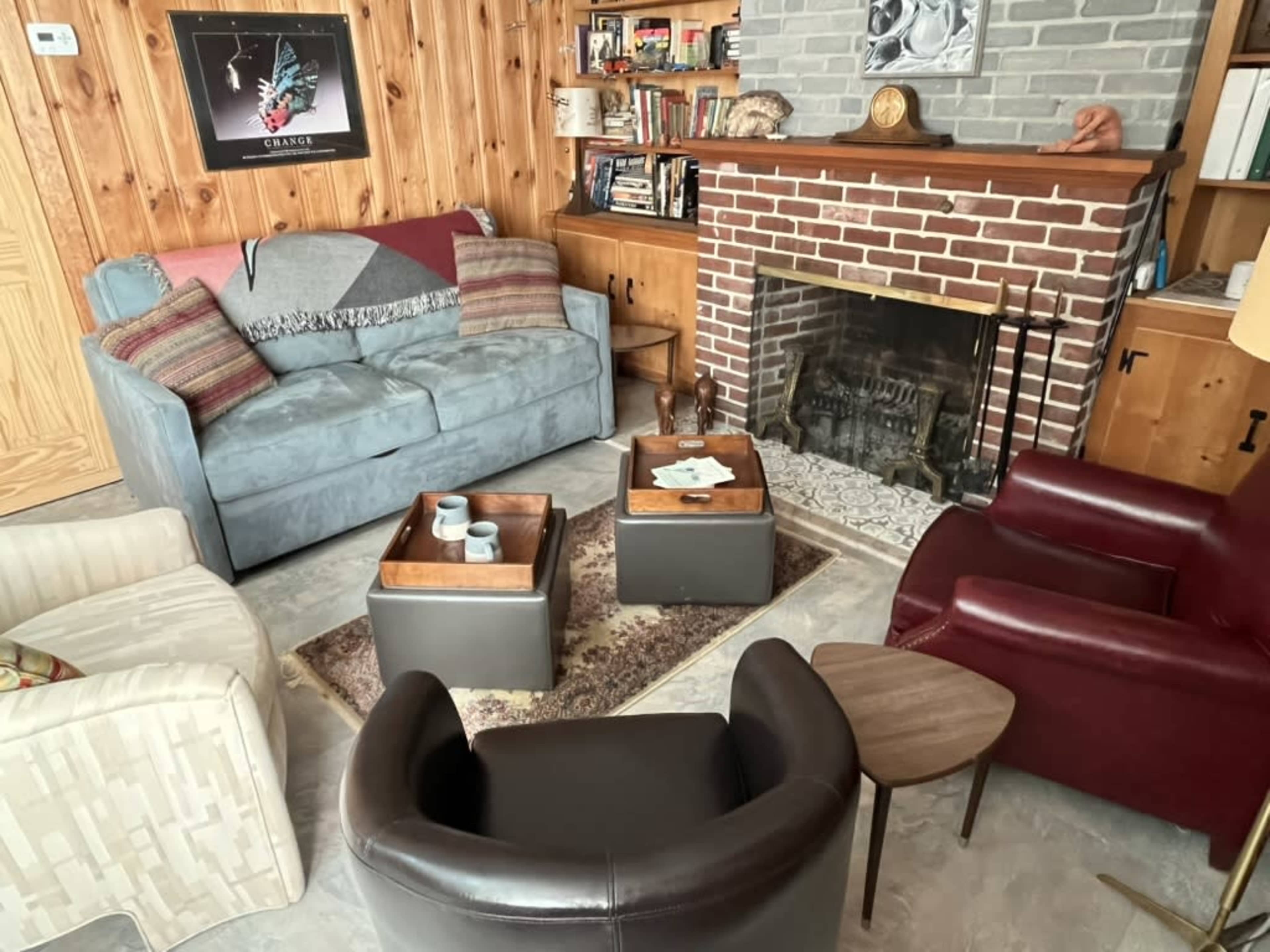 The image shows a cozy living room with a fireplace, featuring a blue couch, a maroon armchair, two small coffee tables, and a wood-paneled wall adorned with books and decorative items.