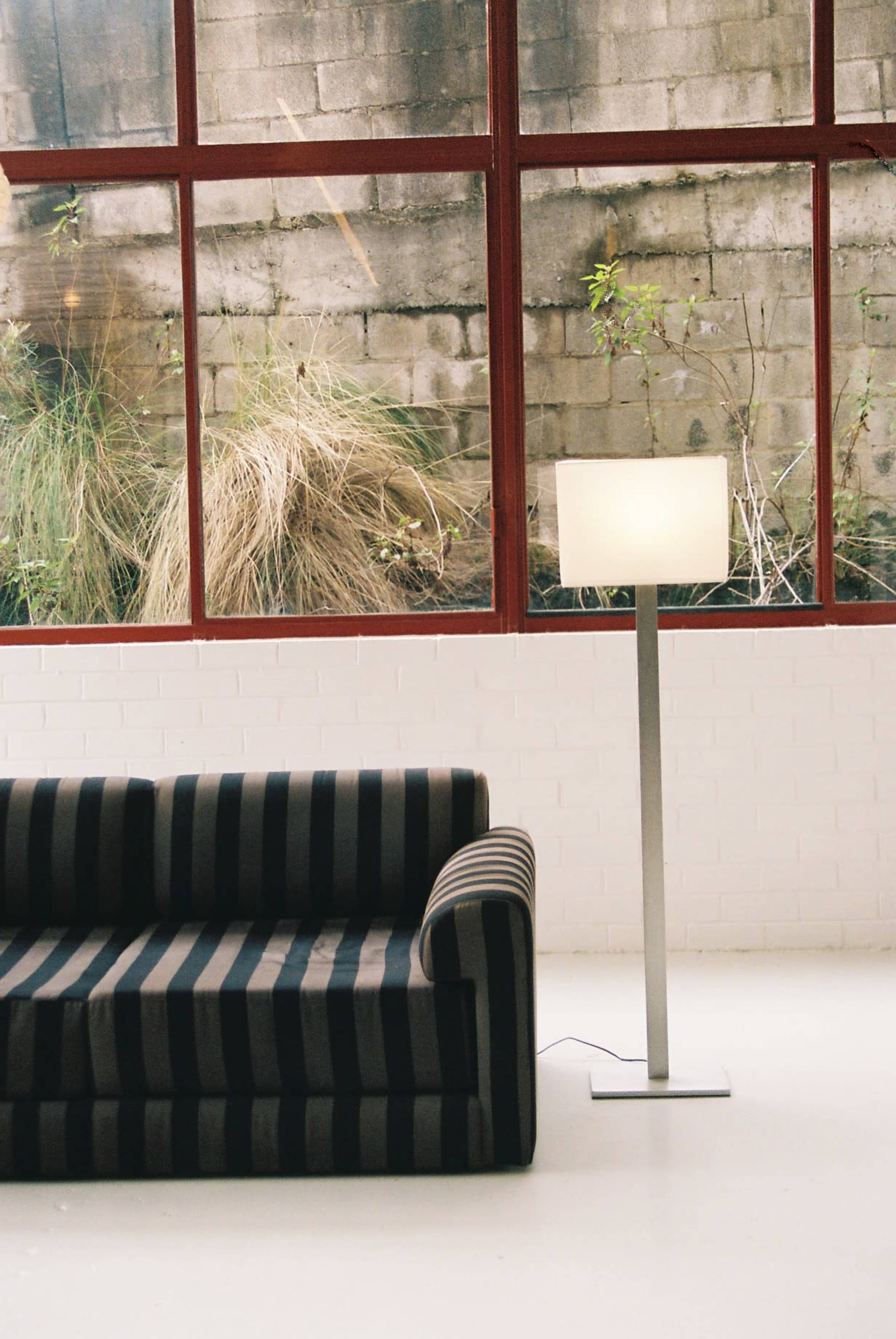The image shows a striped sofa next to a floor lamp in a room with large red-framed windows overlooking a brick wall and greenery.