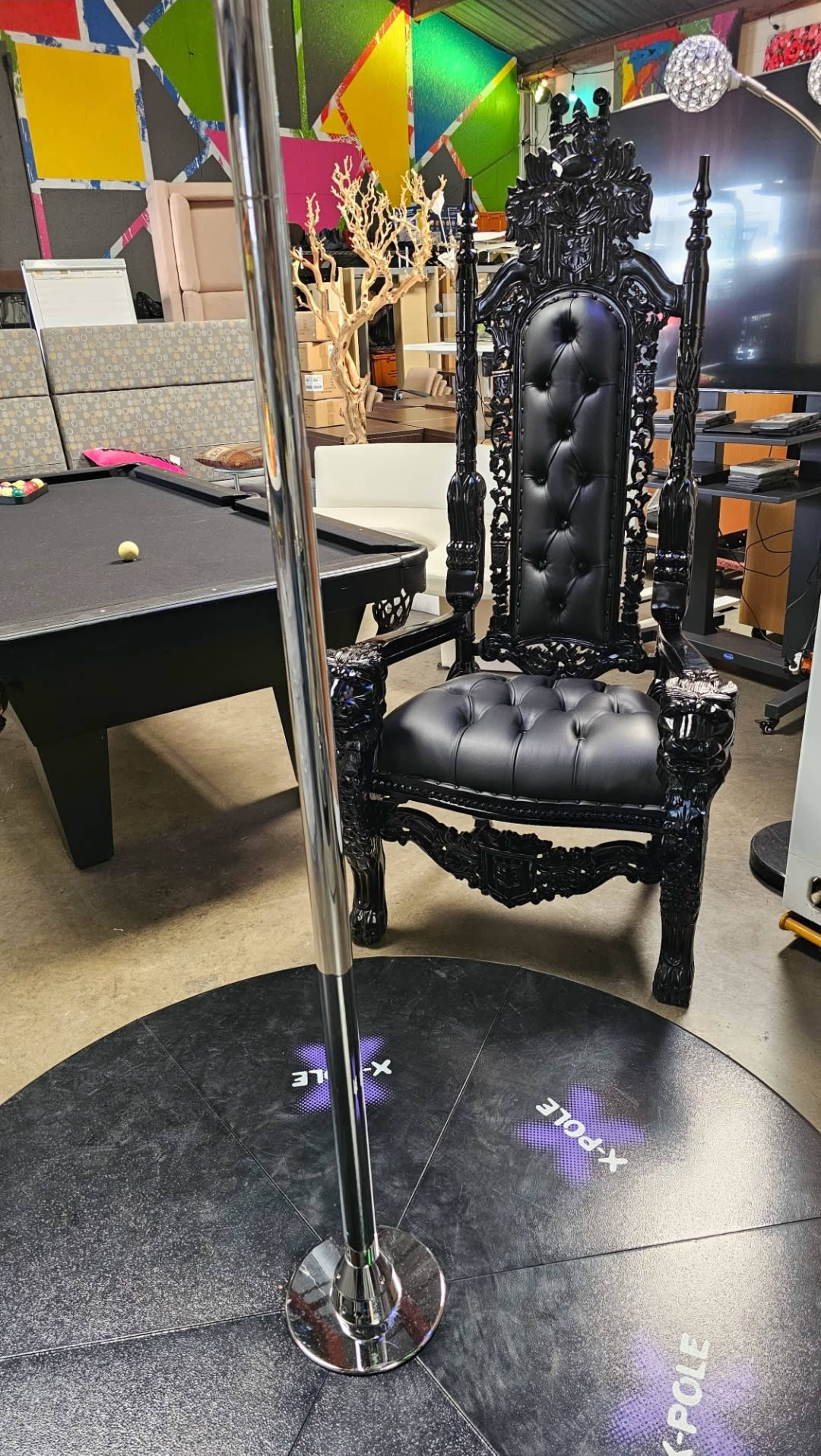 The image shows a shiny chrome pole beside an ornate black chair in a room filled with colorful decor and furniture.