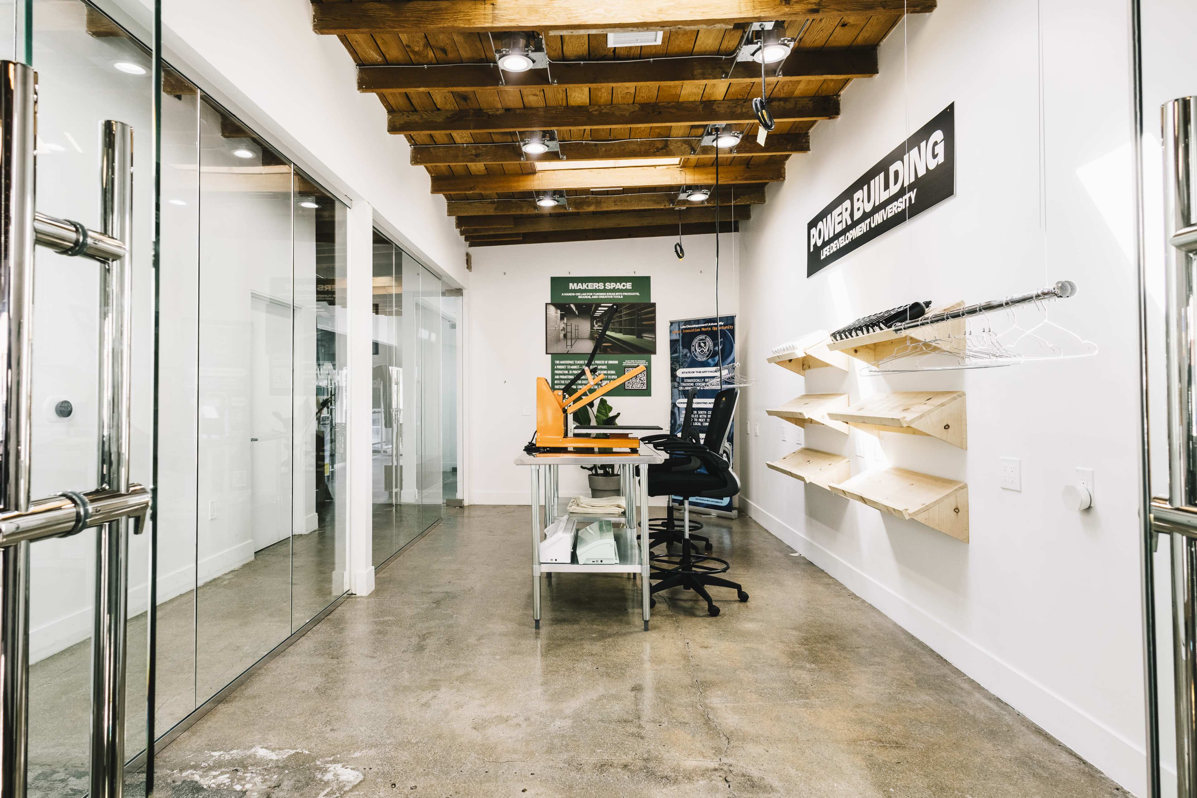 The image shows a modern, minimalist workspace with a clear glass wall, wooden beams on the ceiling, and shelving for display items.