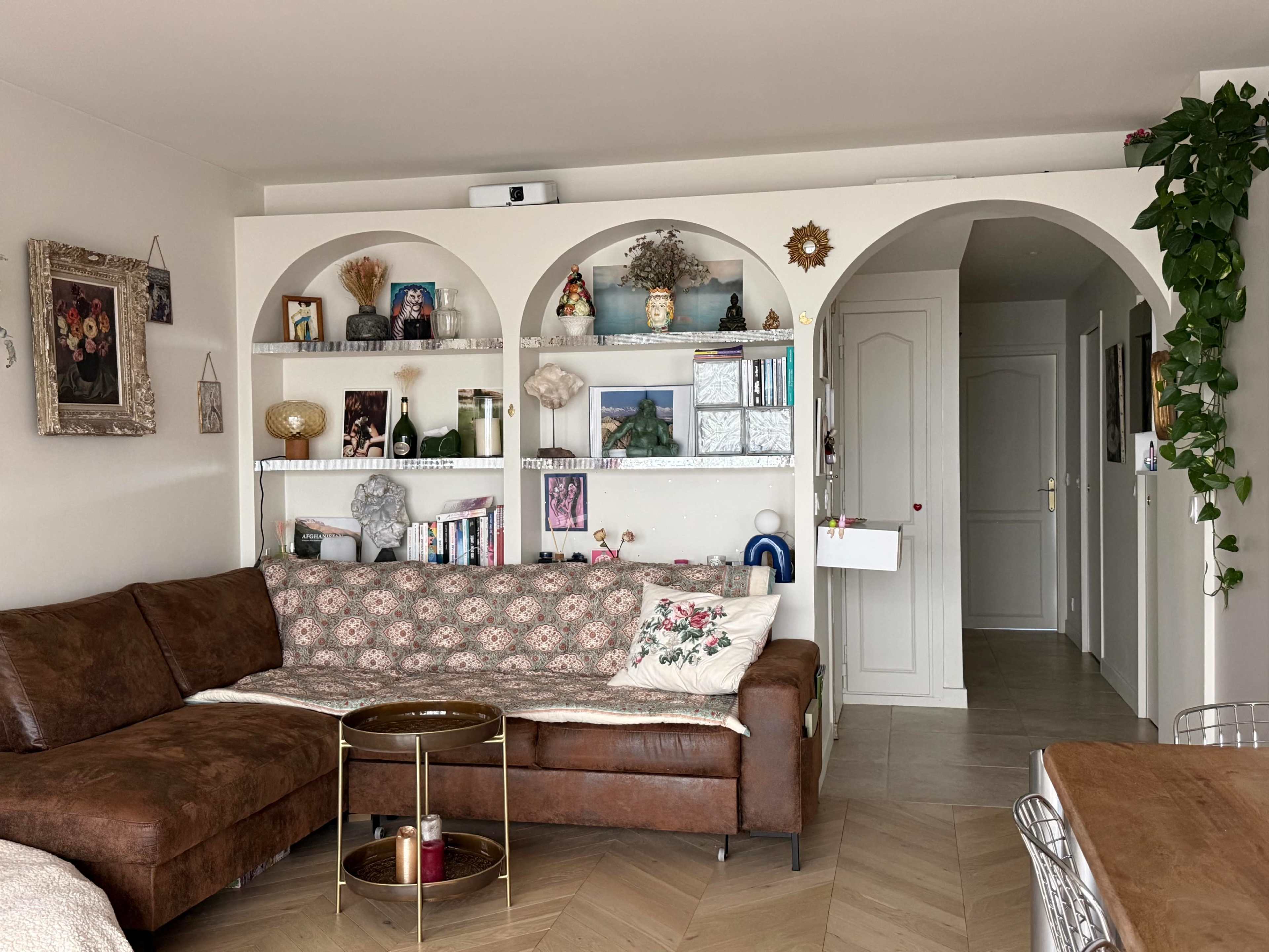 The image shows a cozy living room with a brown sofa, decorative shelves filled with books and ornaments, and an adjoining hallway.