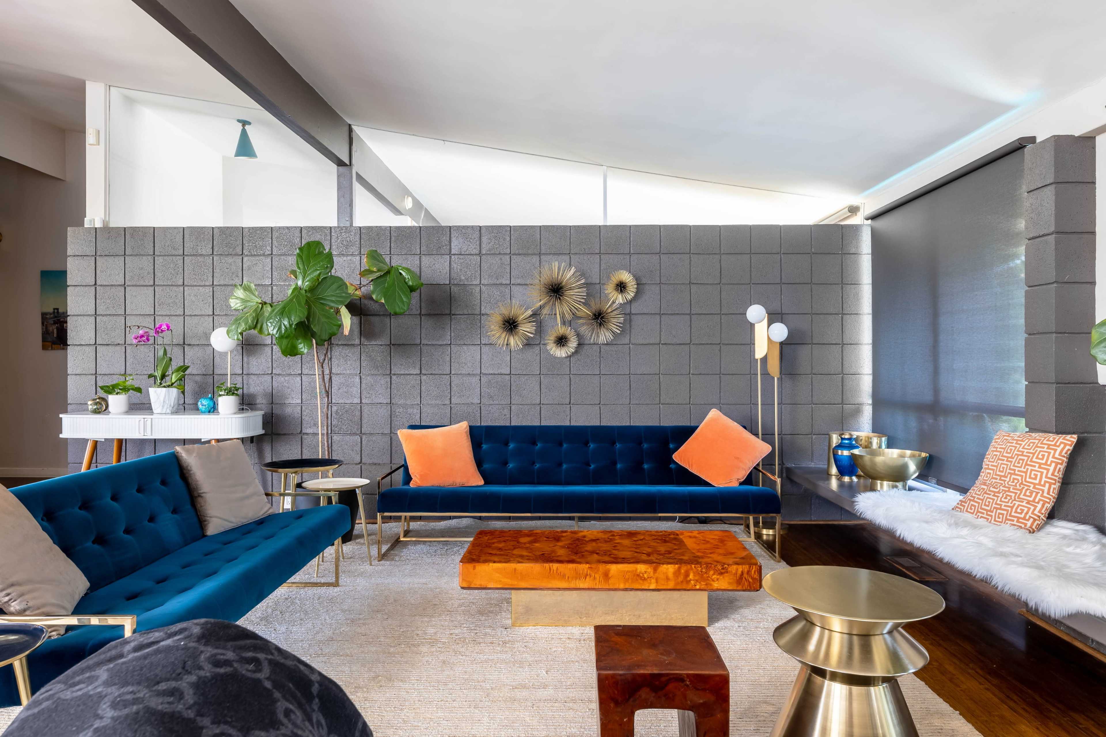 The living room features blue velvet sofas, a wooden coffee table, green plants, and modern lighting against a textured gray wall.