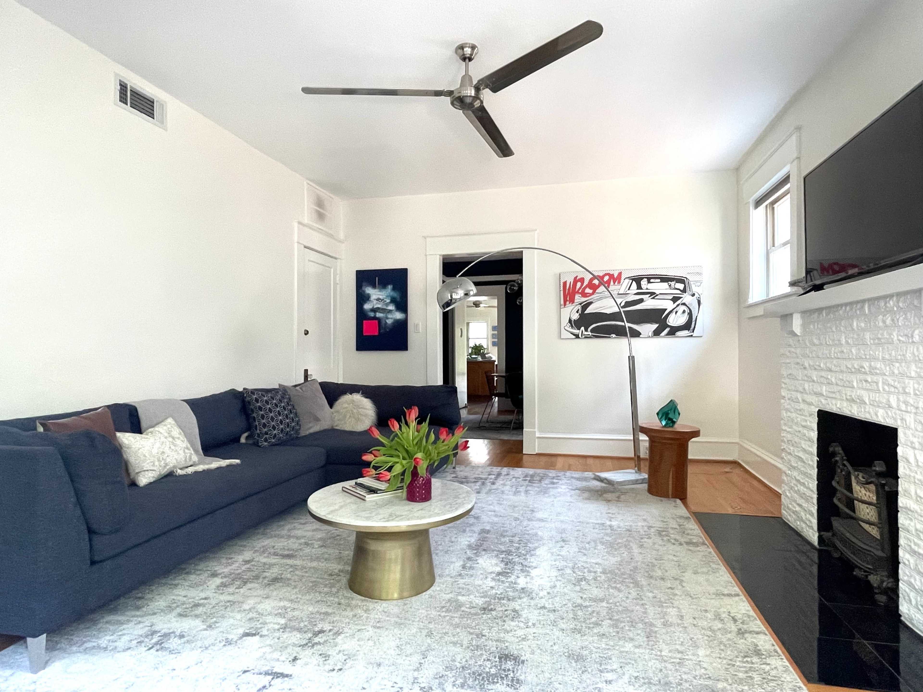 A spacious living room features a dark blue sofa, a round marble coffee table, and a white brick fireplace, with a modern ceiling fan overhead and artwork on the walls.
