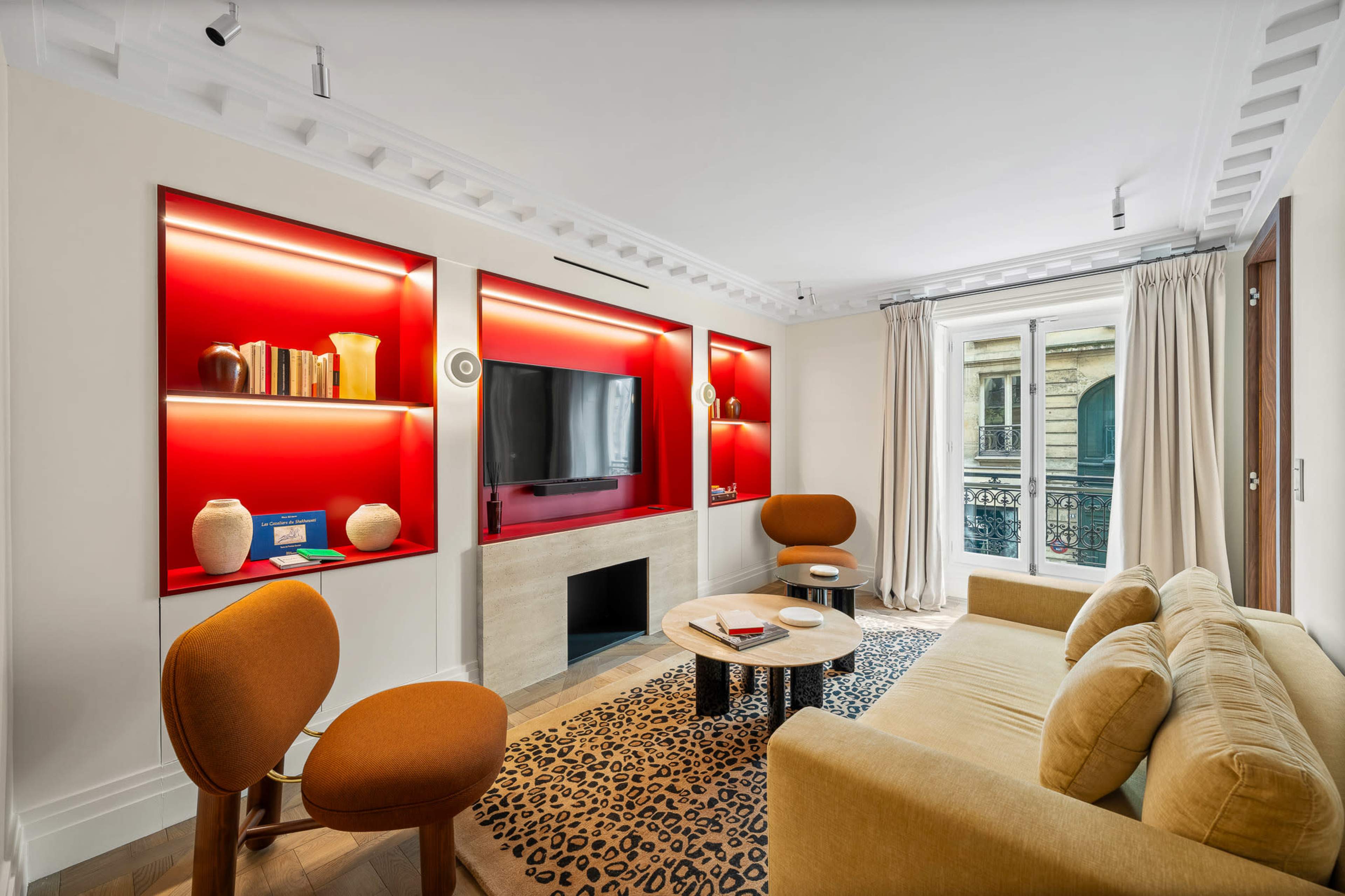 The image shows a modern living room with a beige sofa, orange accent chairs, a circular coffee table, and a wall-mounted television surrounded by red-lit shelves.
