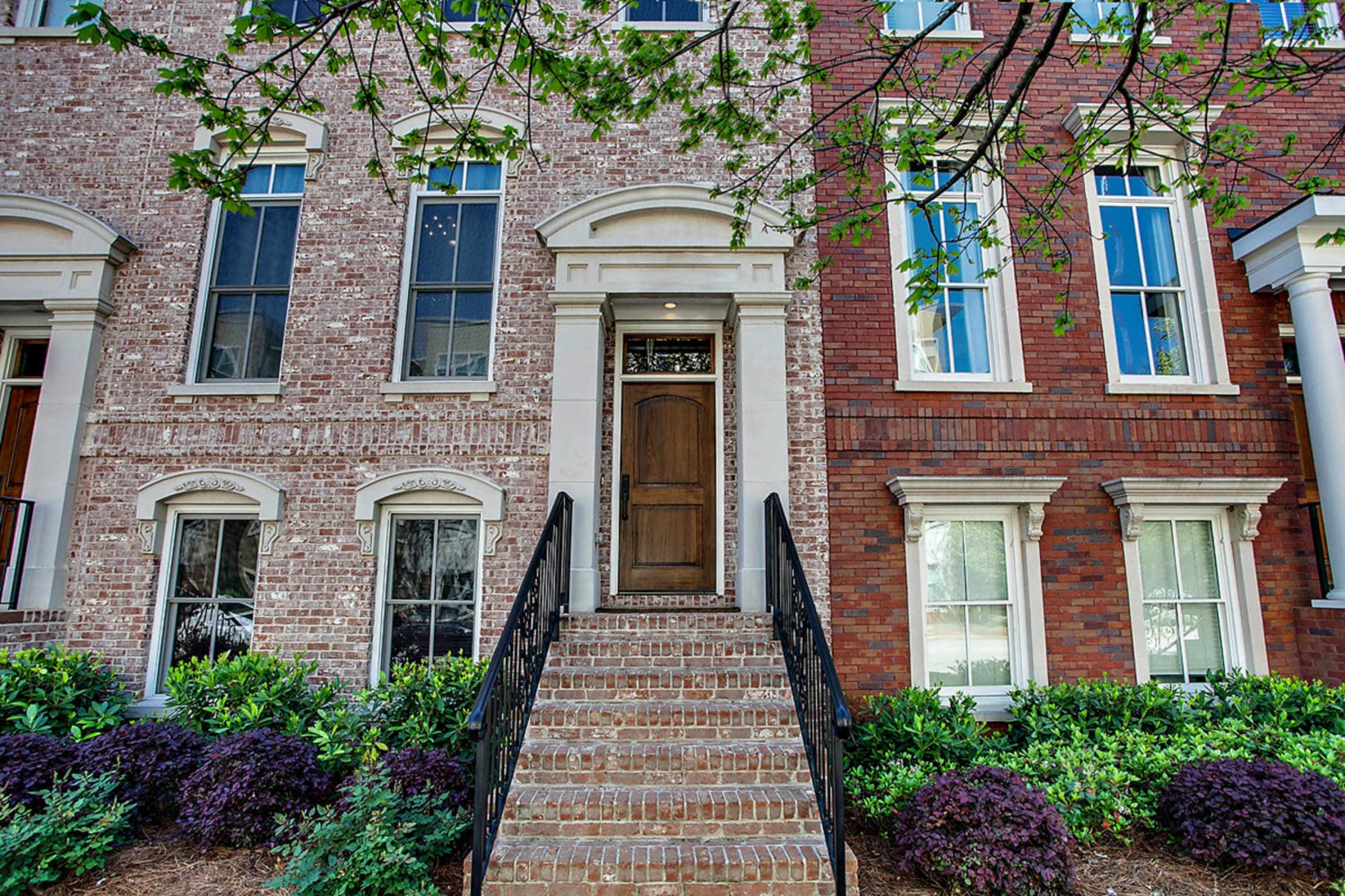 The image shows a brick townhouse with a central wooden door, flanked by tall windows and flanked by brick and greenery on either side.