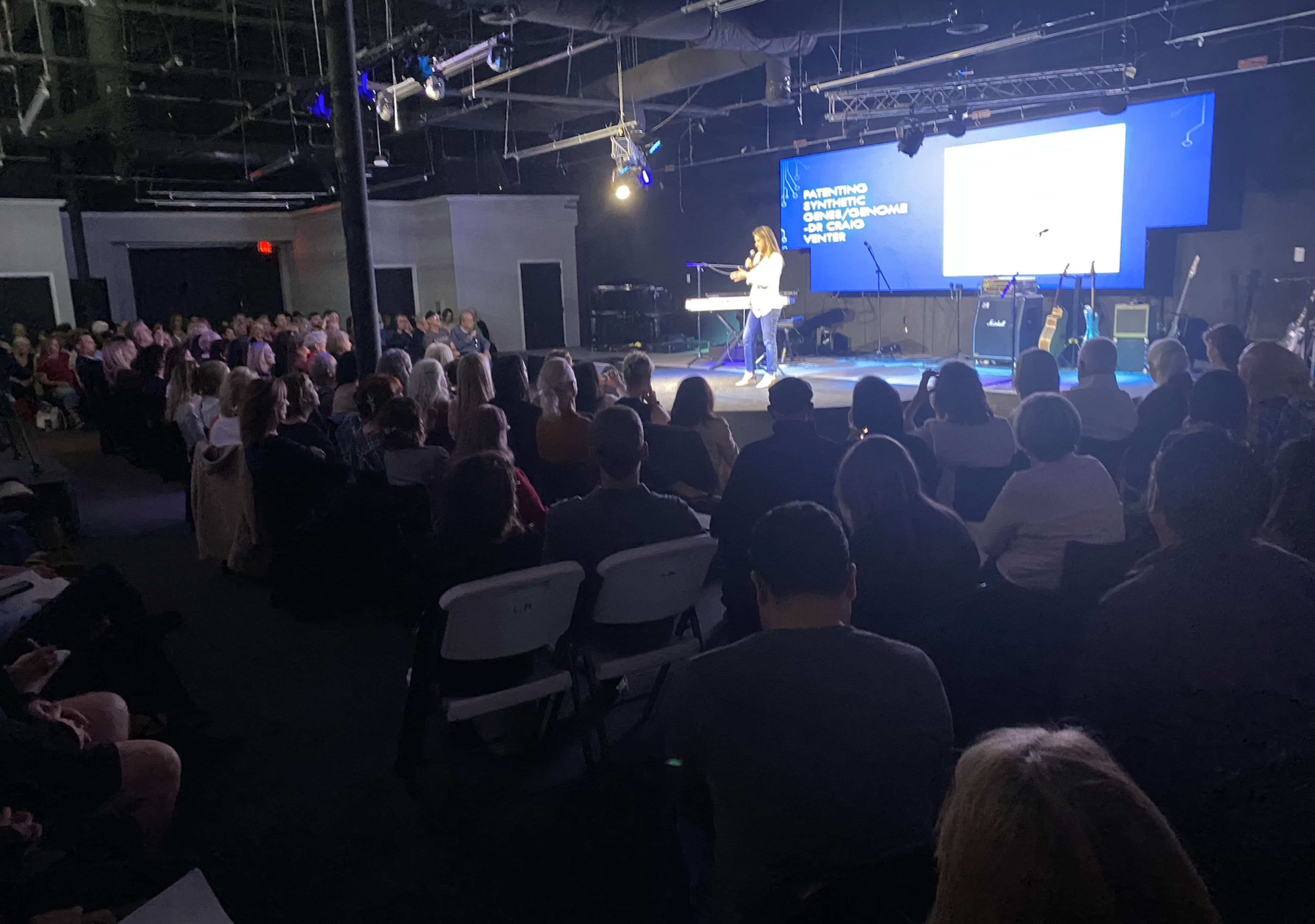 A speaker presents to a seated audience in a dimly lit venue with stage lighting and a large screen.