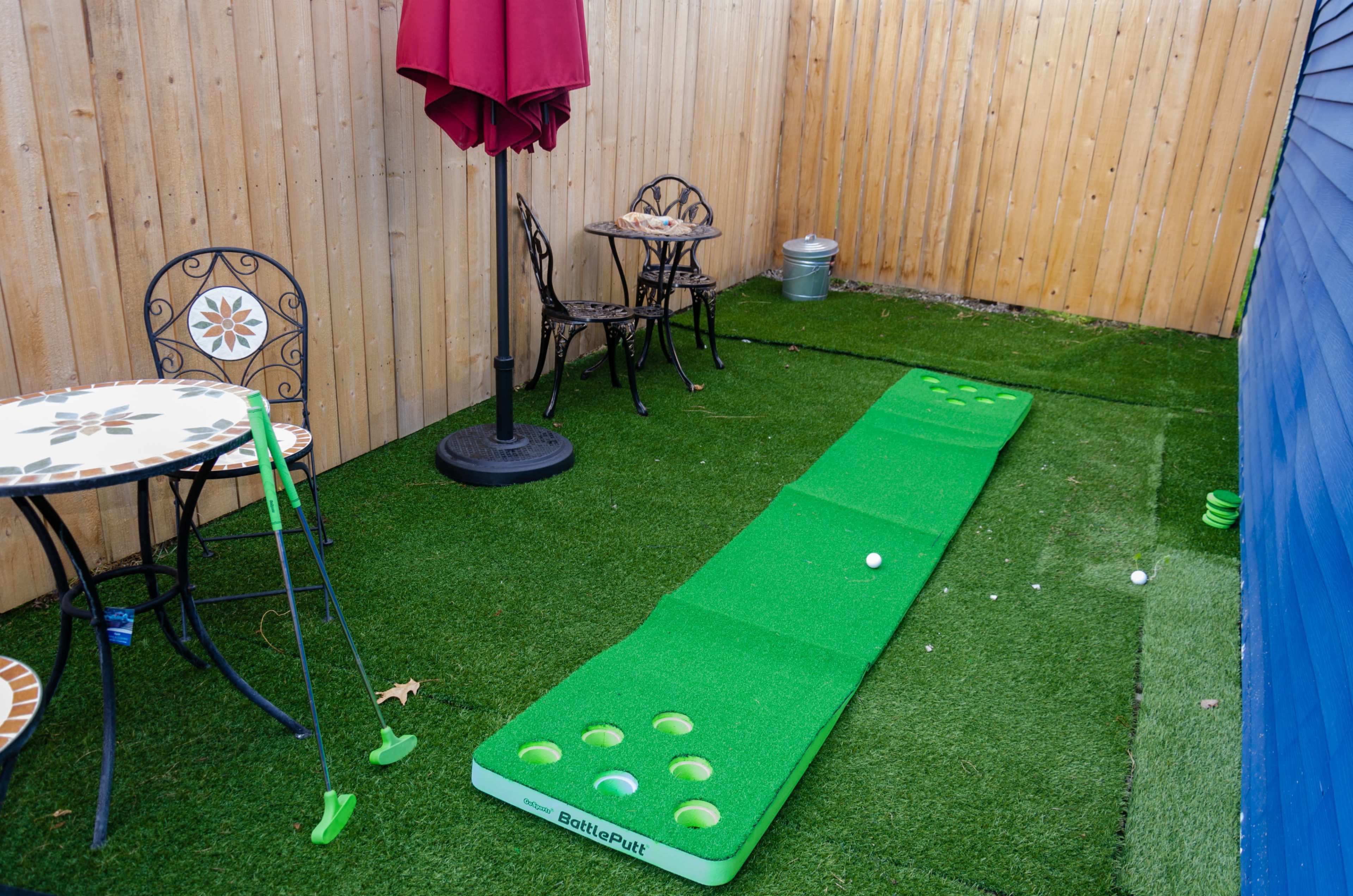 A small outdoor area with a putting green, featuring a golf ball near a series of holes, surrounded by chairs and a table beneath a red umbrella.