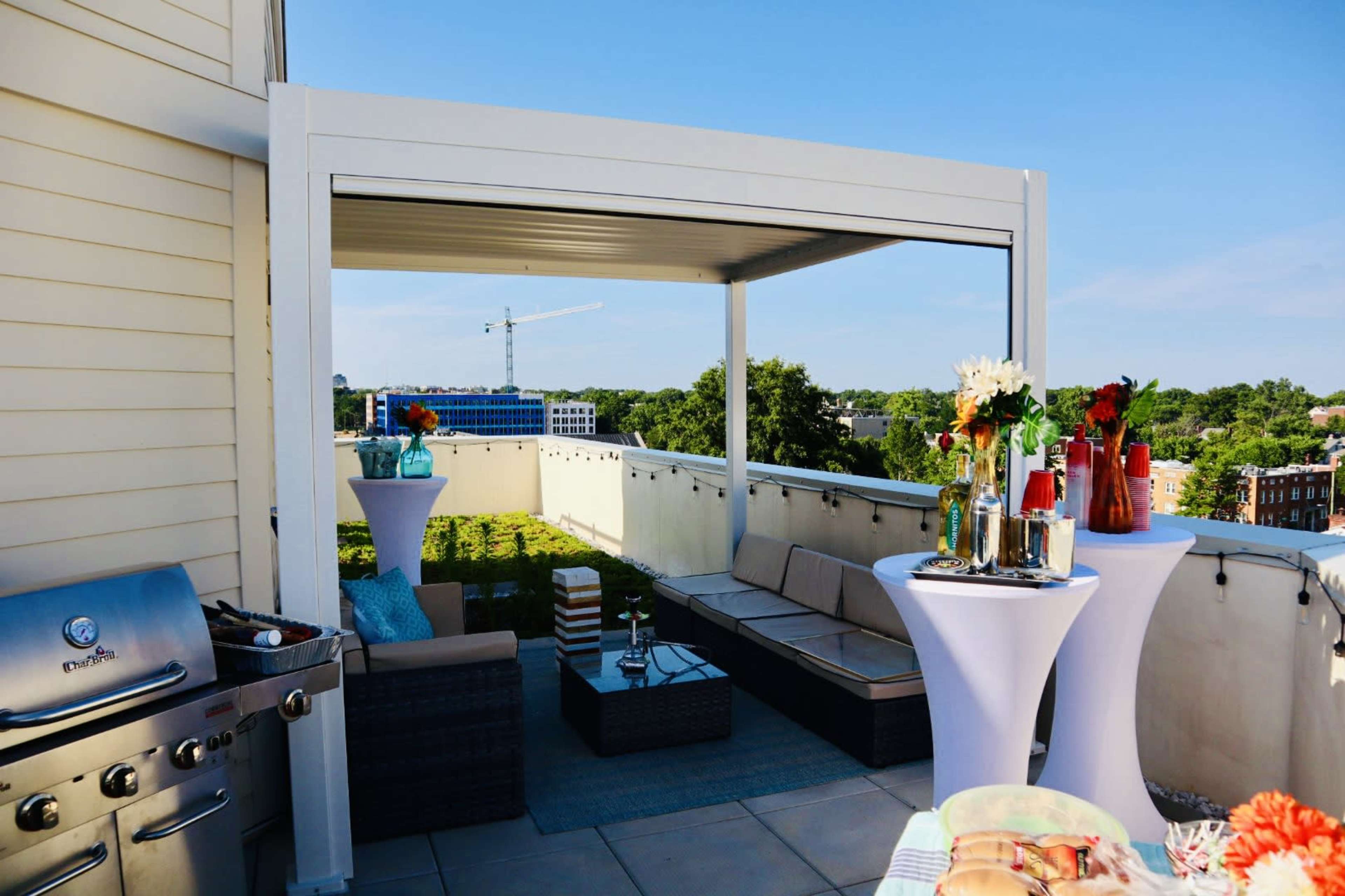 A rooftop patio features a seating area with a sectional sofa, a grill, and two high tables adorned with drinks and flowers, overlooking a construction site in the distance.