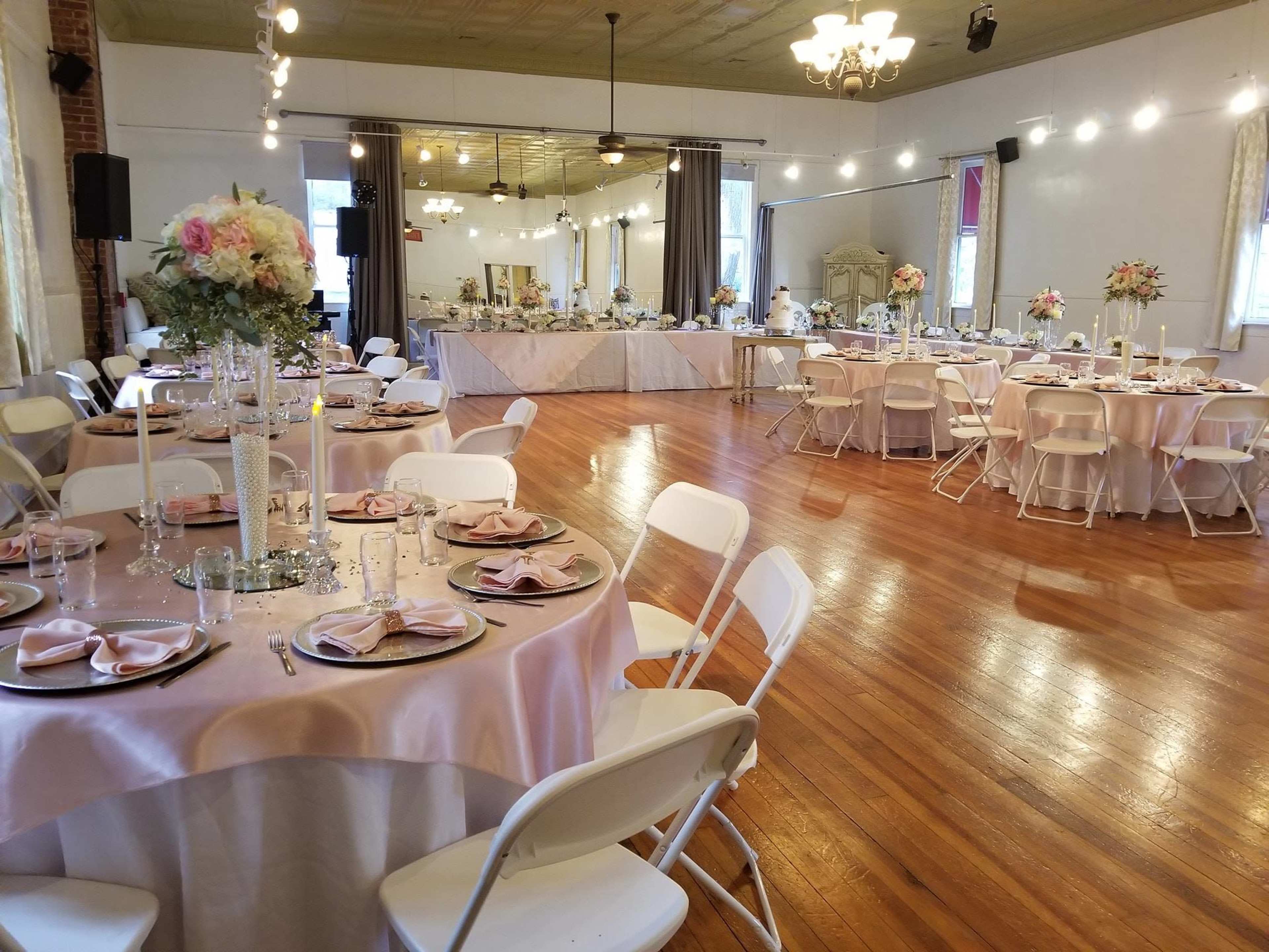The image shows a banquet hall set up for an event, featuring round tables with pink tablecloths and white chairs, adorned with floral centerpieces and candles, alongside a buffet table in the background.