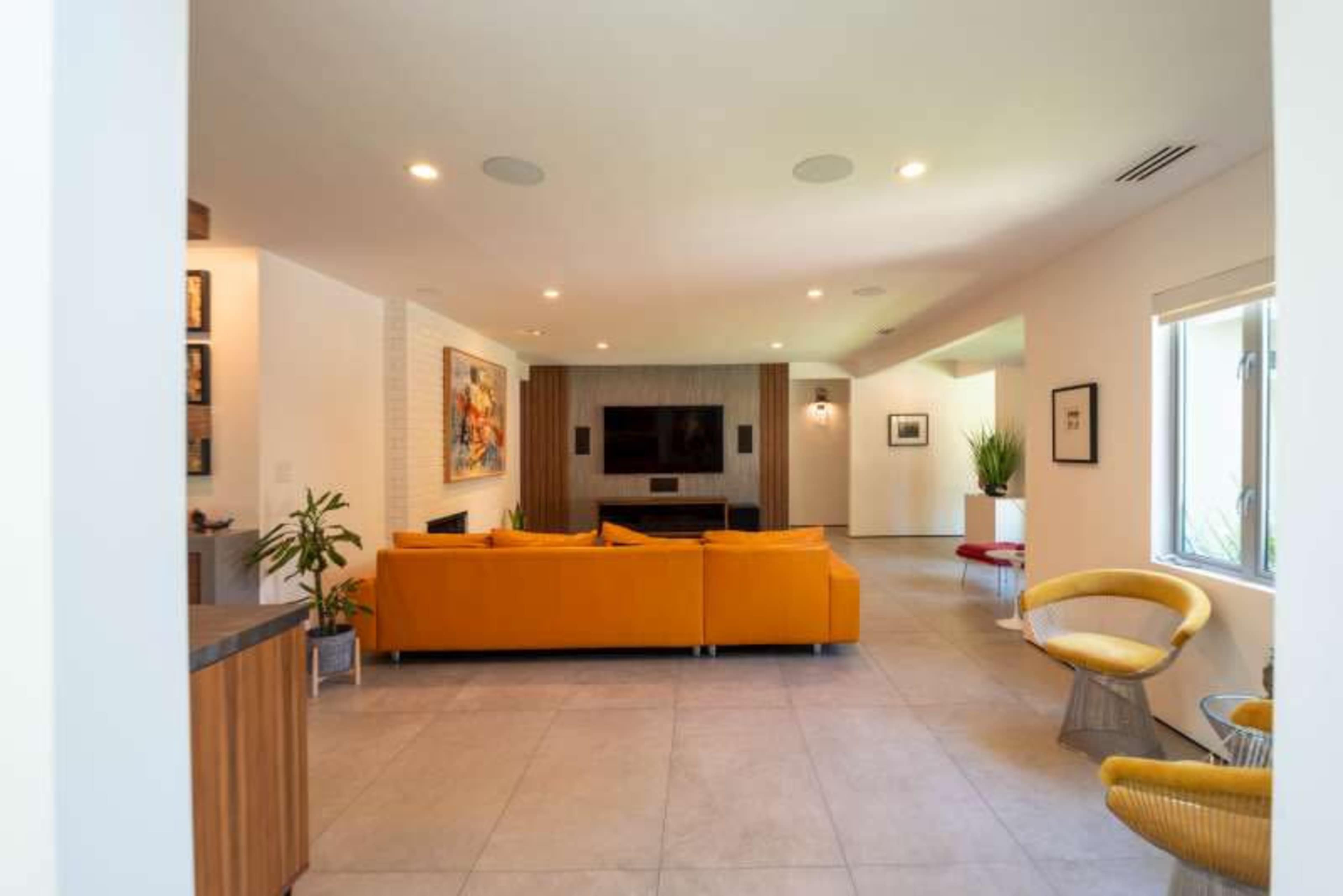 A spacious living room features a yellow sofa, a flat-screen TV mounted on a wooden wall, and windows allowing natural light.