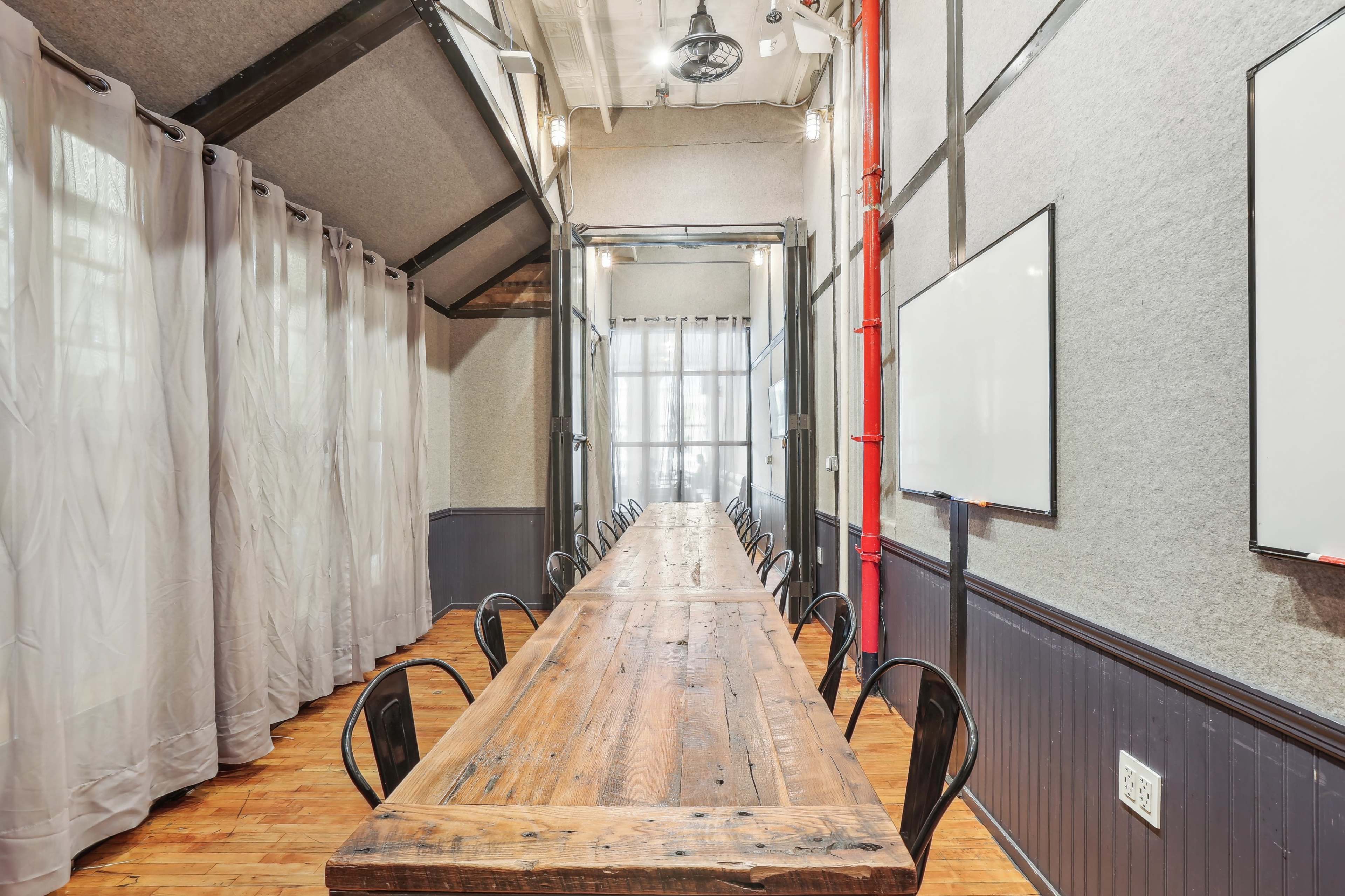 A long wooden table is arranged in a well-lit meeting room with gray walls and large windows, surrounded by black chairs.