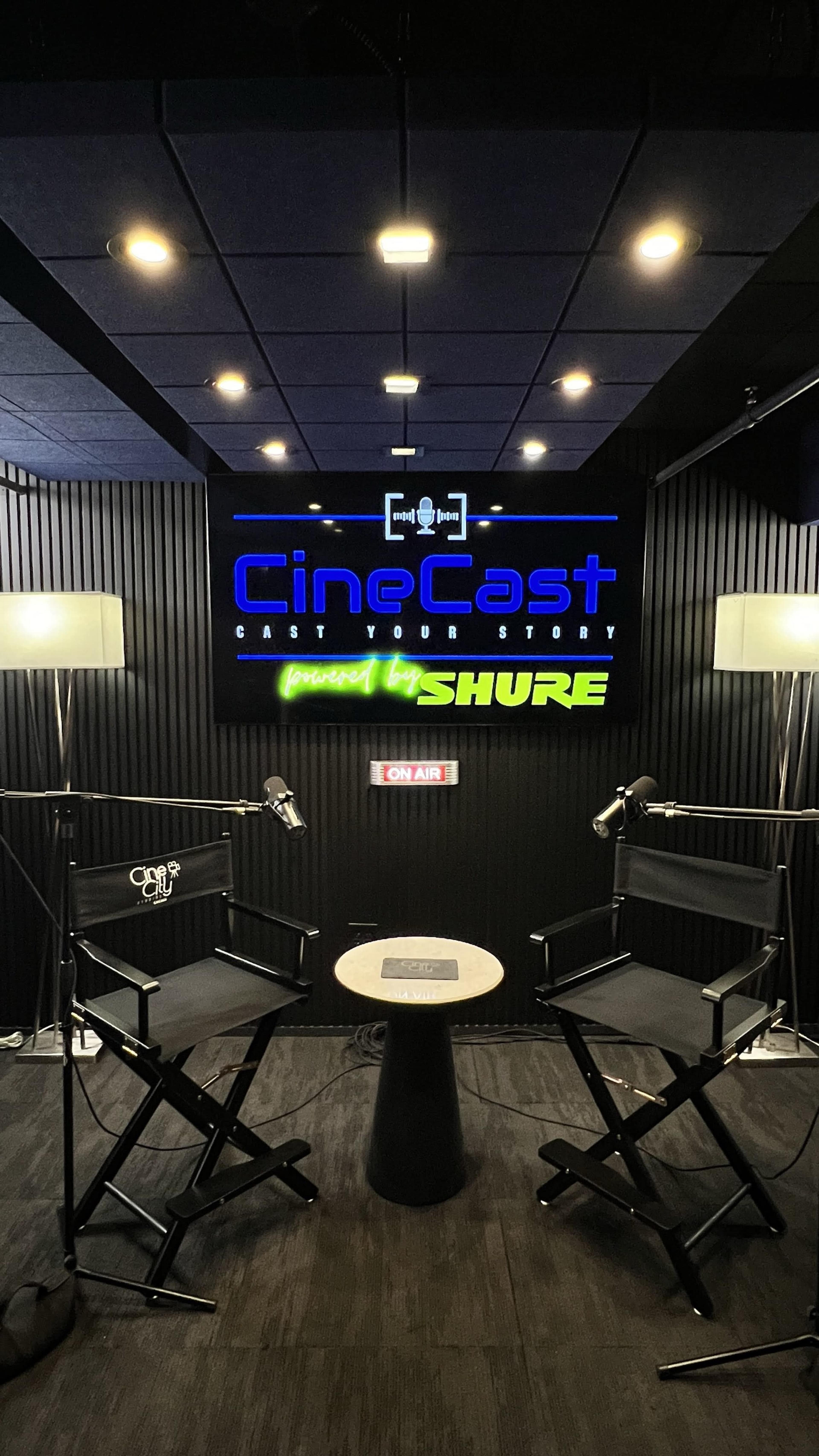 The image shows a modern podcast studio featuring two director's chairs, a small table in the center, and a large screen displaying the "CineCast" logo, all under bright ceiling lights.