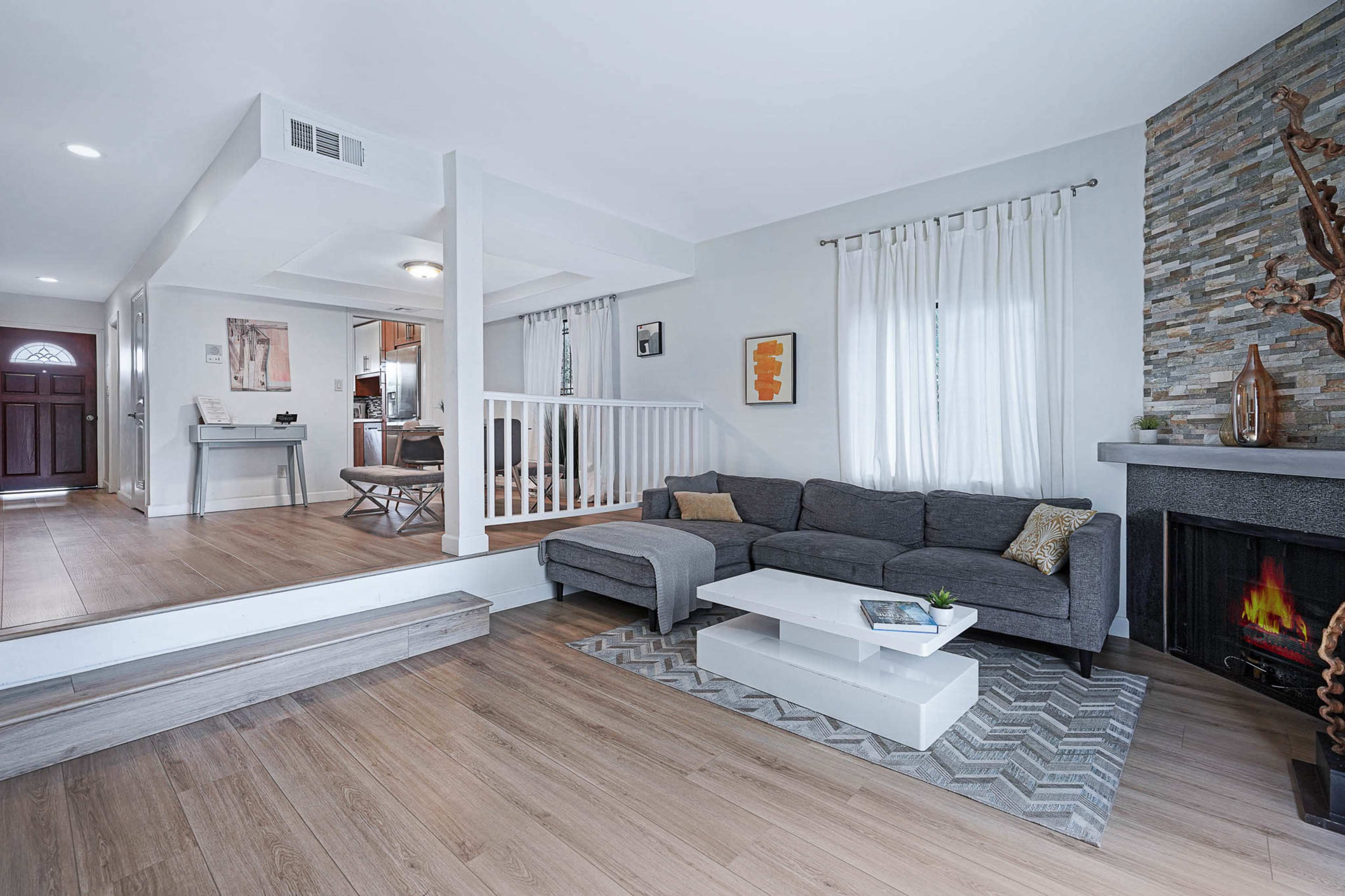 The image shows a modern living room with a gray sectional sofa, a white coffee table on a geometric rug, and a stone fireplace, illuminated by natural light coming through sheer curtains.