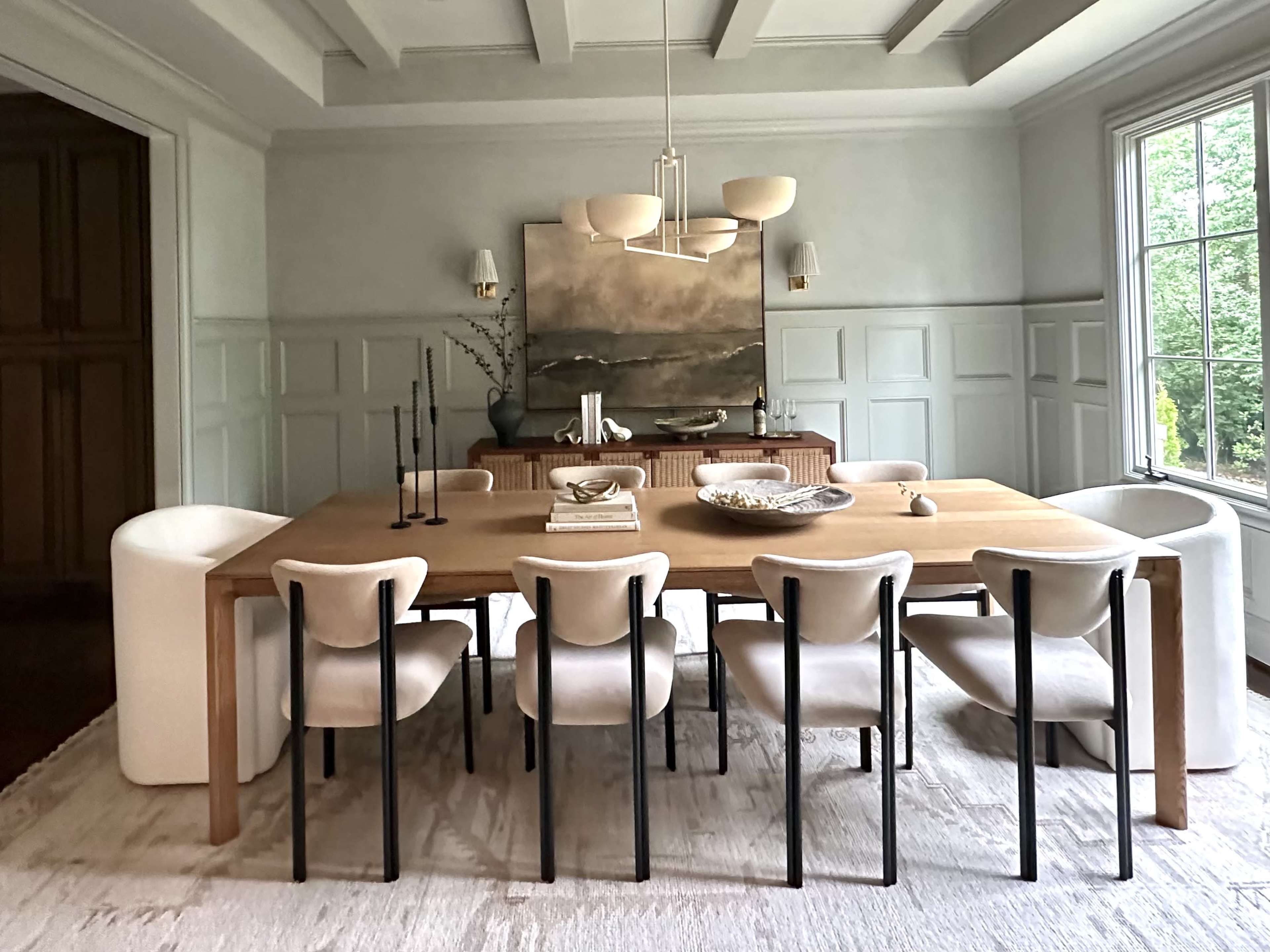 The image shows a modern dining room featuring a large wooden table surrounded by eight upholstered chairs, with a decorative centerpiece and a large artwork on the wall.