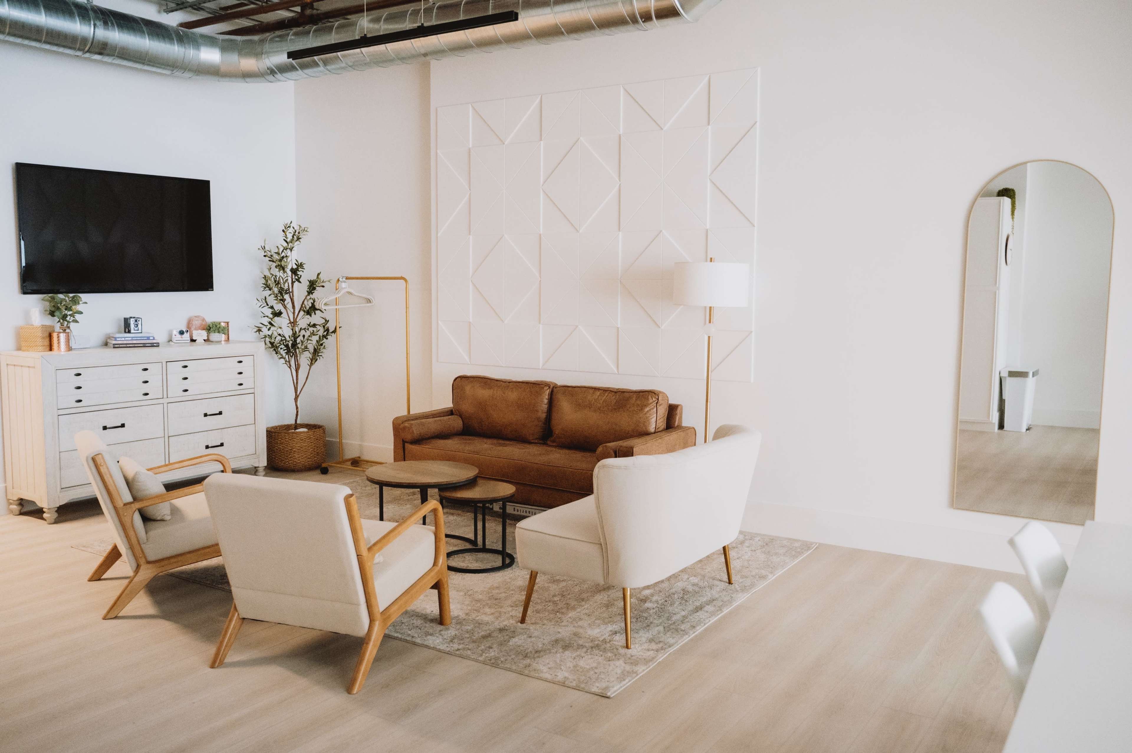 A modern living room features a brown leather sofa, two white chairs, a coffee table, a large mirror, and a TV mounted on the wall.