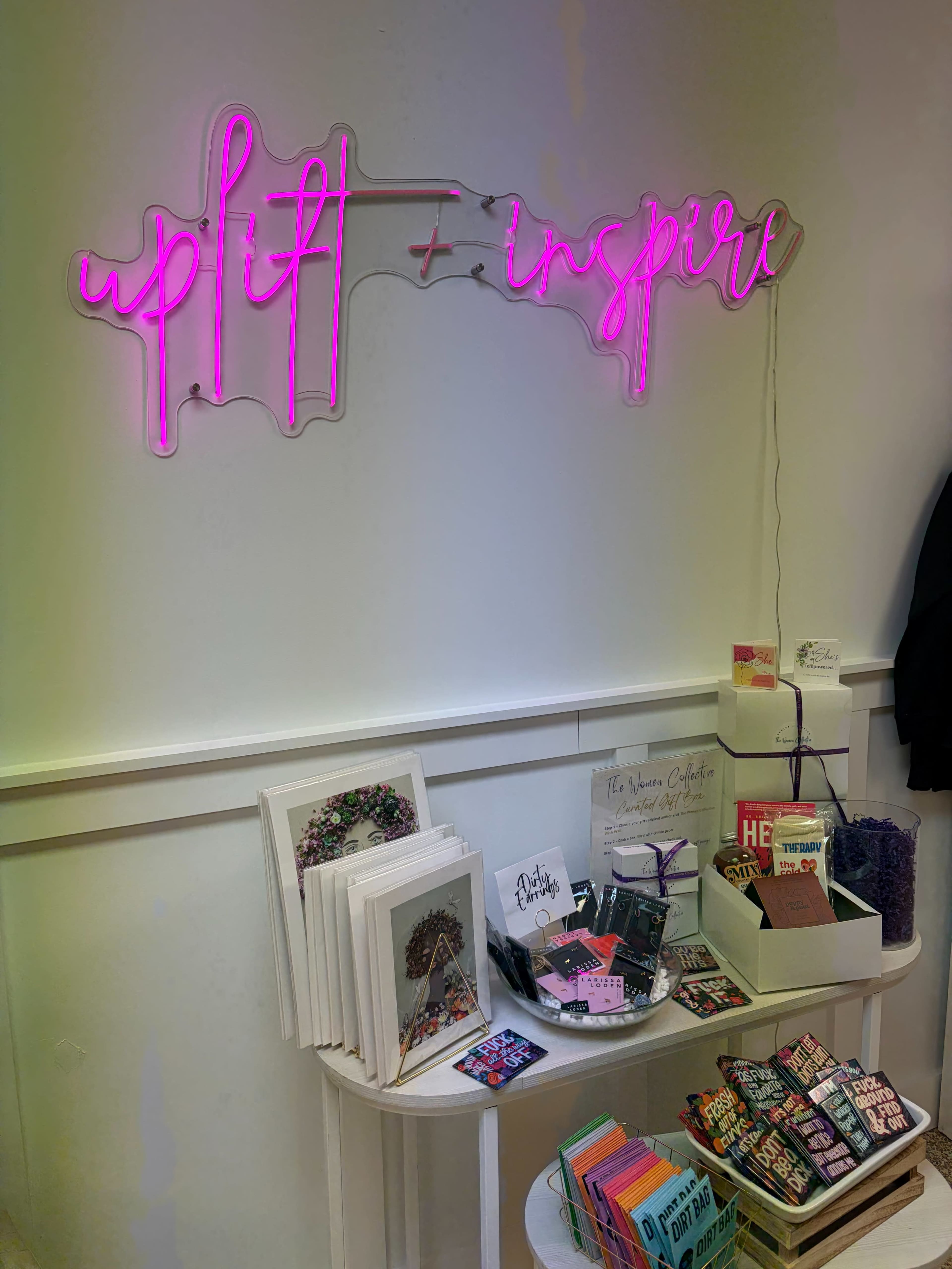A neon sign saying "uplift + inspire" is displayed on a wall above a table filled with various cards and decorative items.