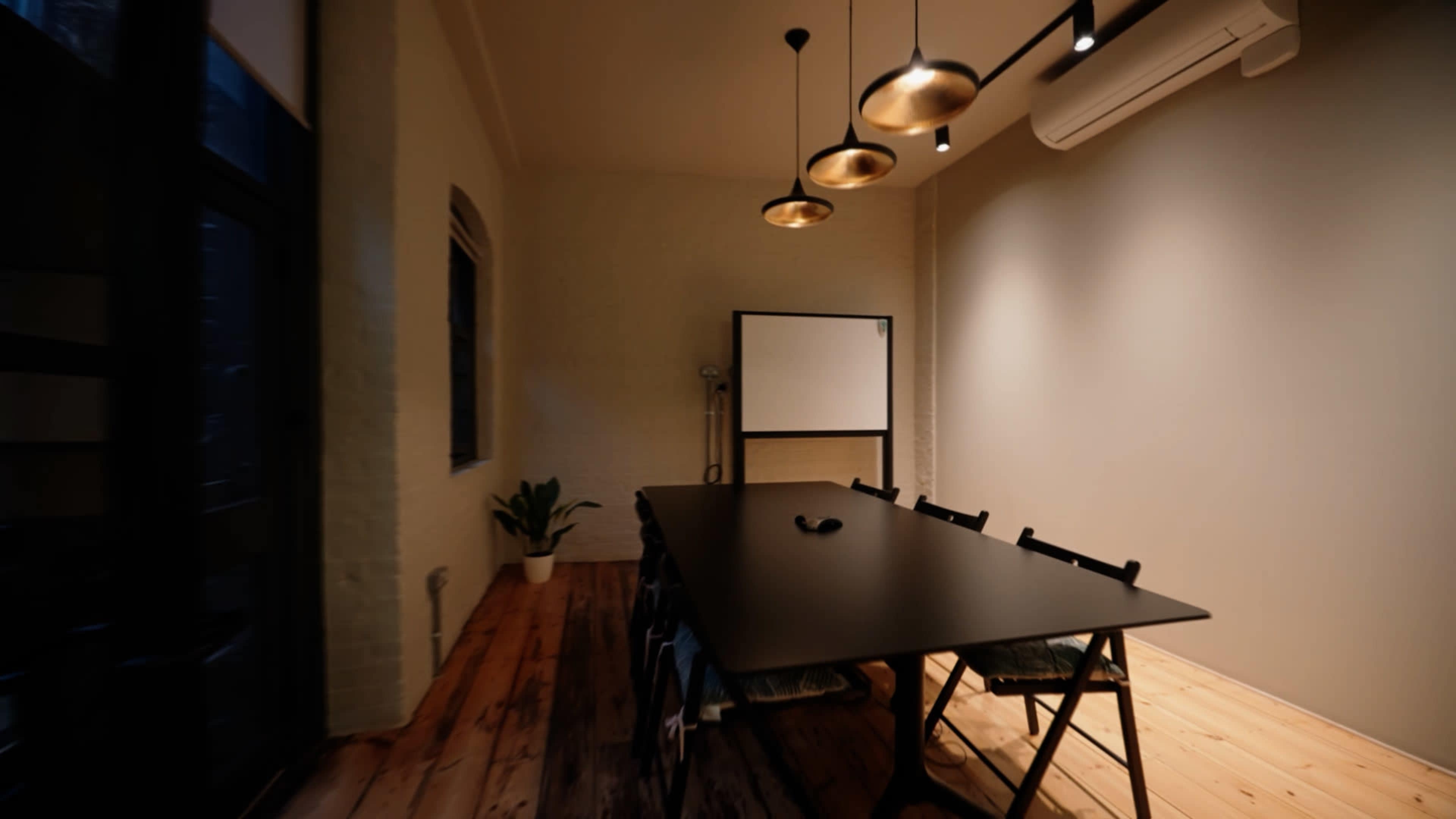 A conference room features a long black table with several chairs, pendant lights overhead, and a blank presentation board against a light-colored wall.