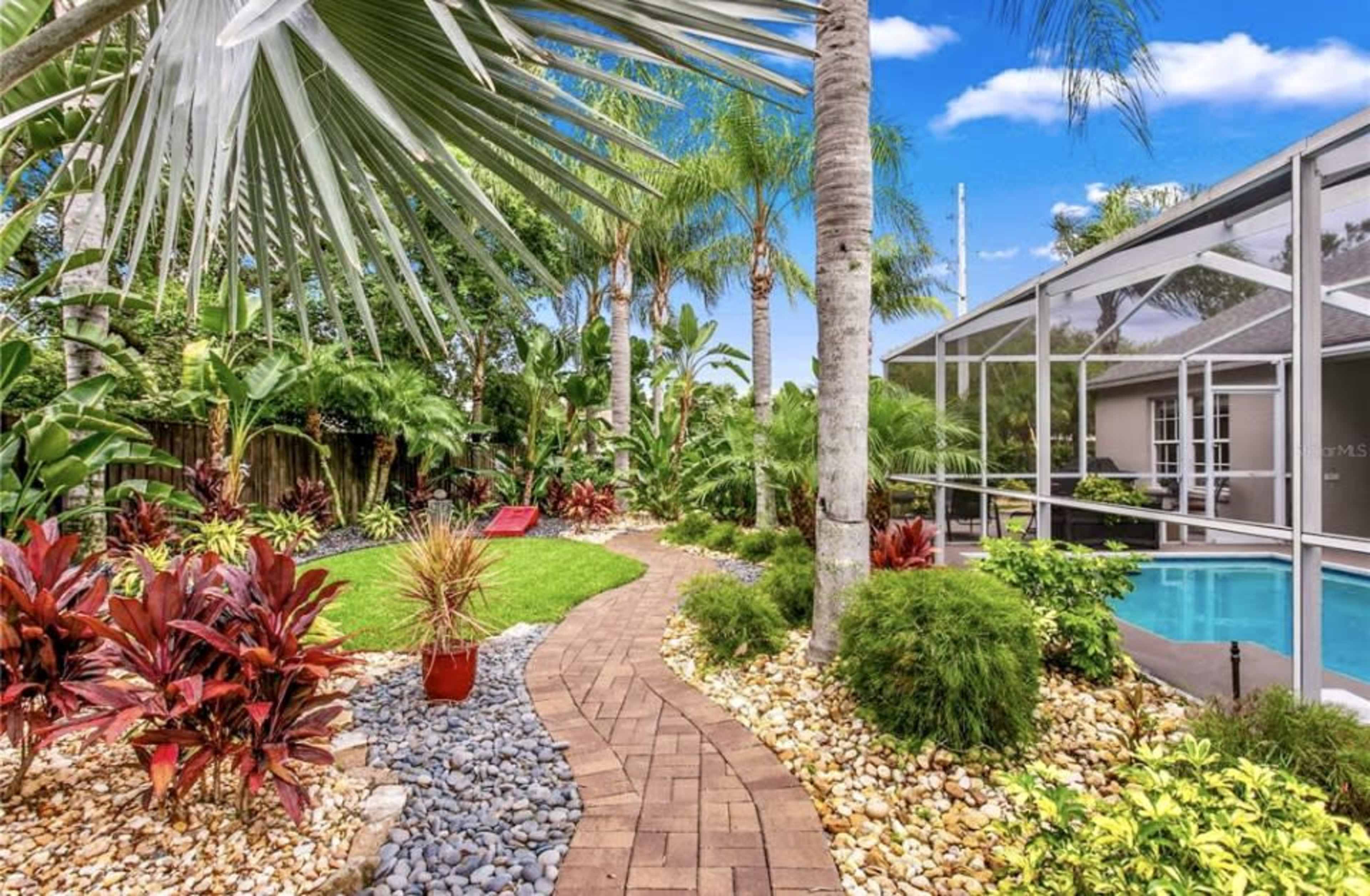 A winding brick pathway leads through a lush garden with tropical plants and a swimming pool in the background.