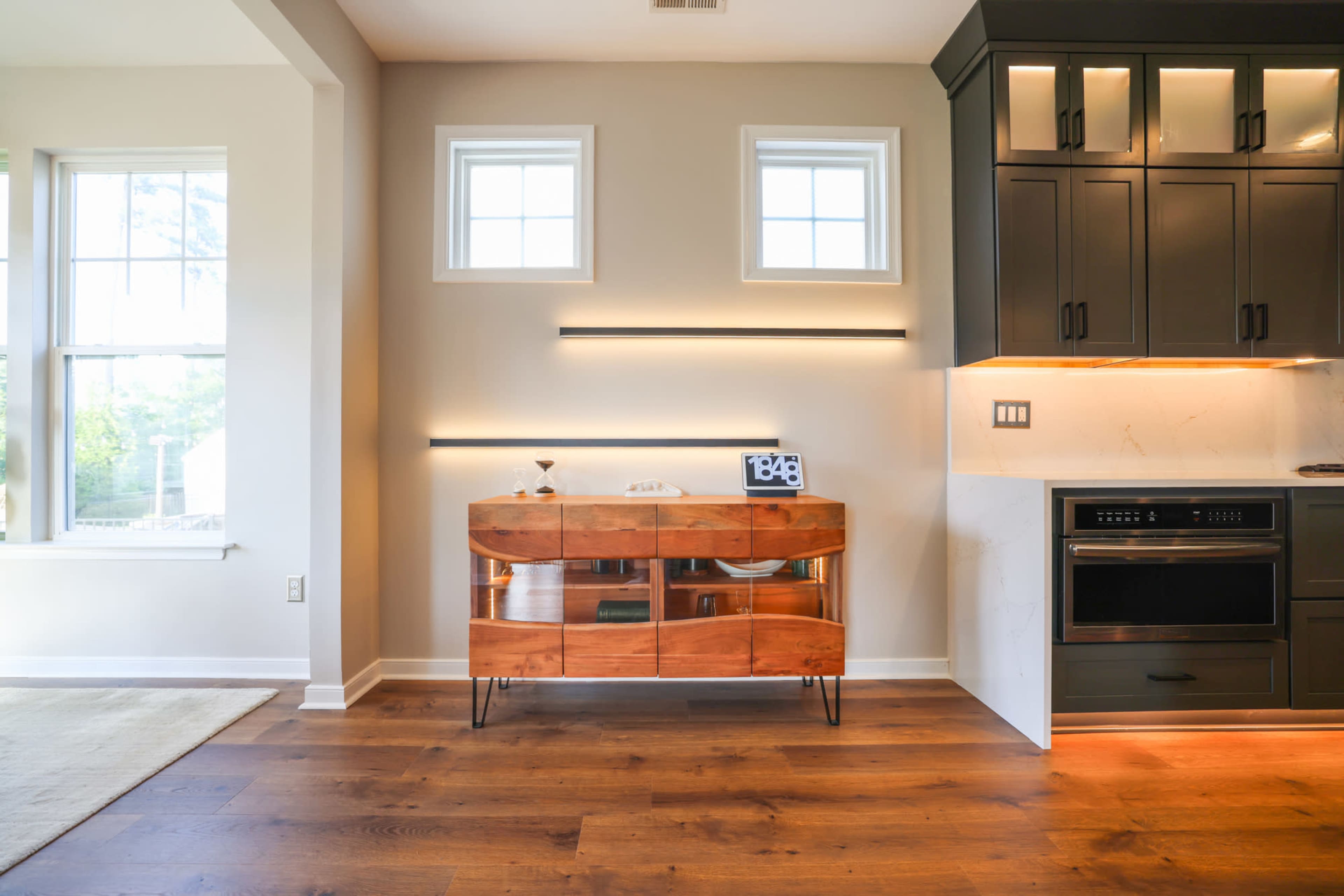 The image shows a modern kitchen area featuring a wooden sideboard against a light-colored wall, with two small windows and sleek upper cabinetry.
