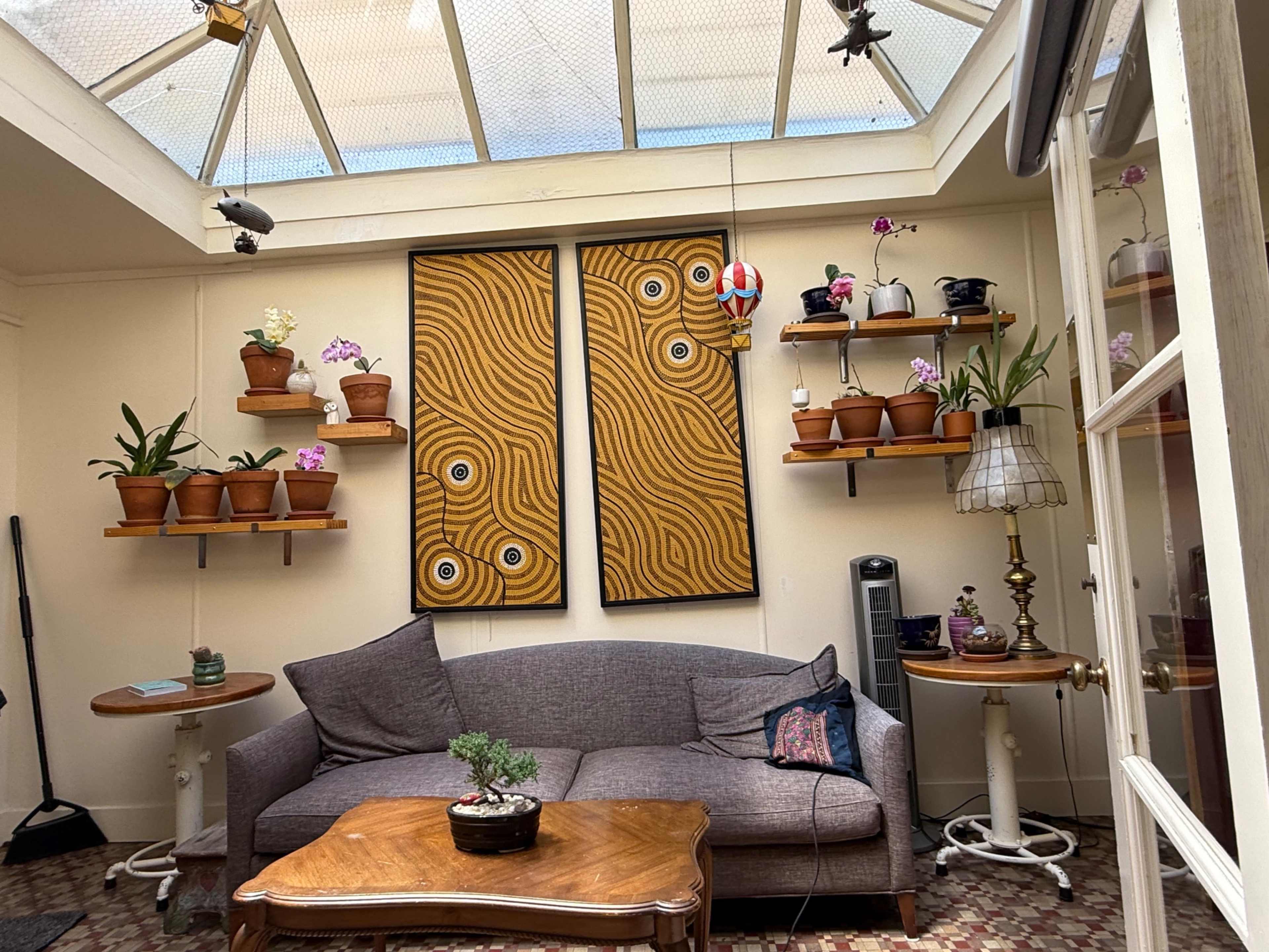 The image shows a cozy indoor space featuring a gray couch, wooden coffee table, and multiple potted plants on shelves, with two large abstract paintings on the wall.