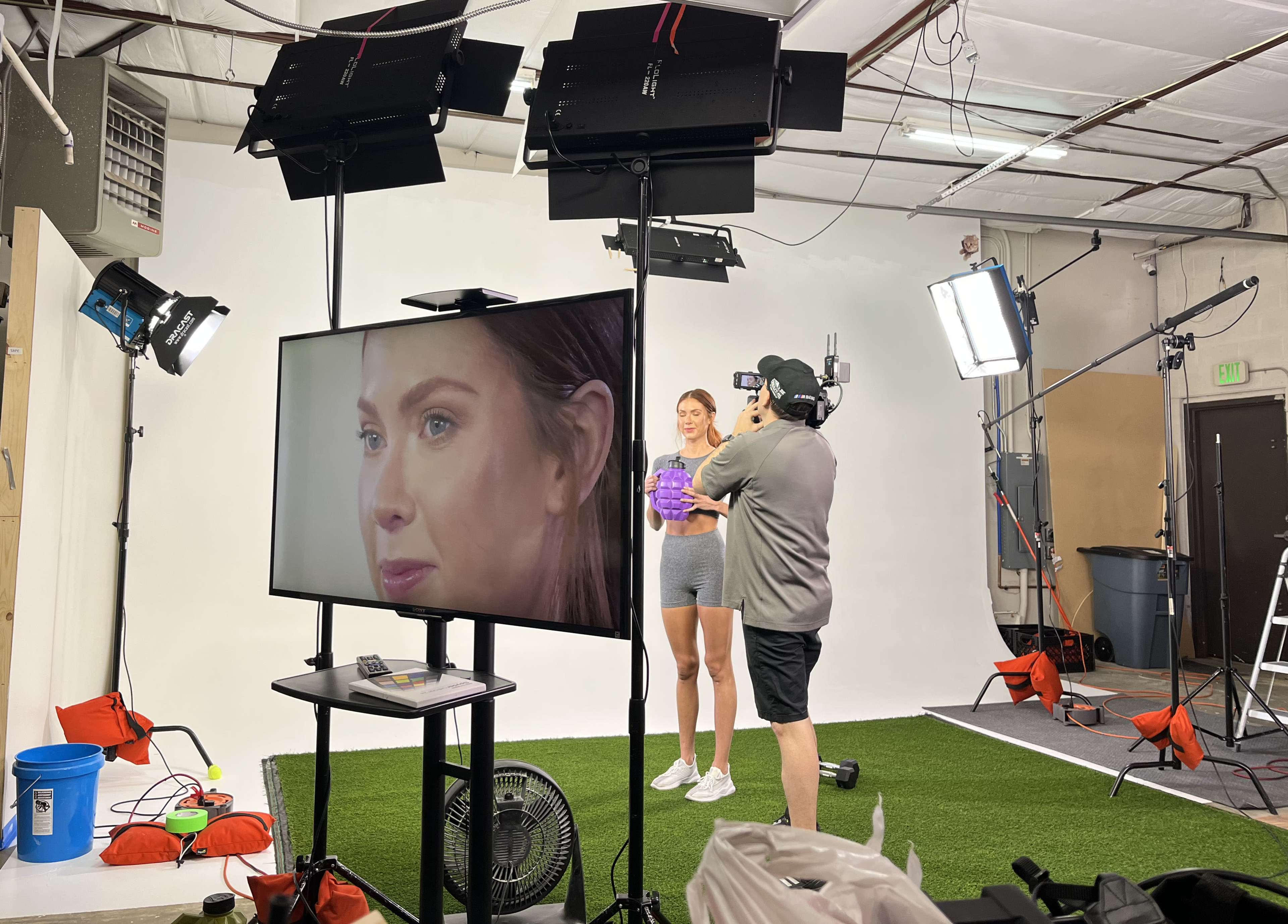 A woman stands in a studio holding a purple object while a man operates a camera and monitors her performance on a screen.