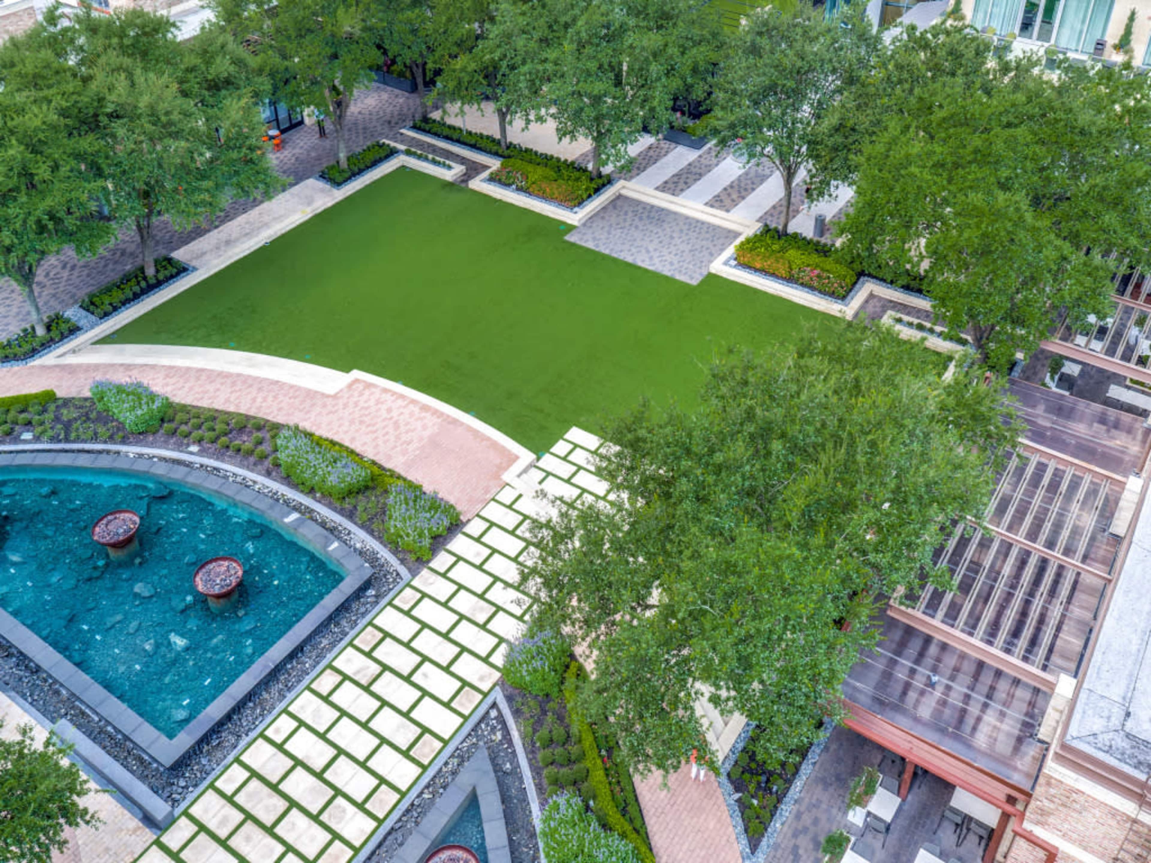 An aerial view of a landscaped outdoor area featuring green grass, stone pathways, a water feature, and surrounding trees.