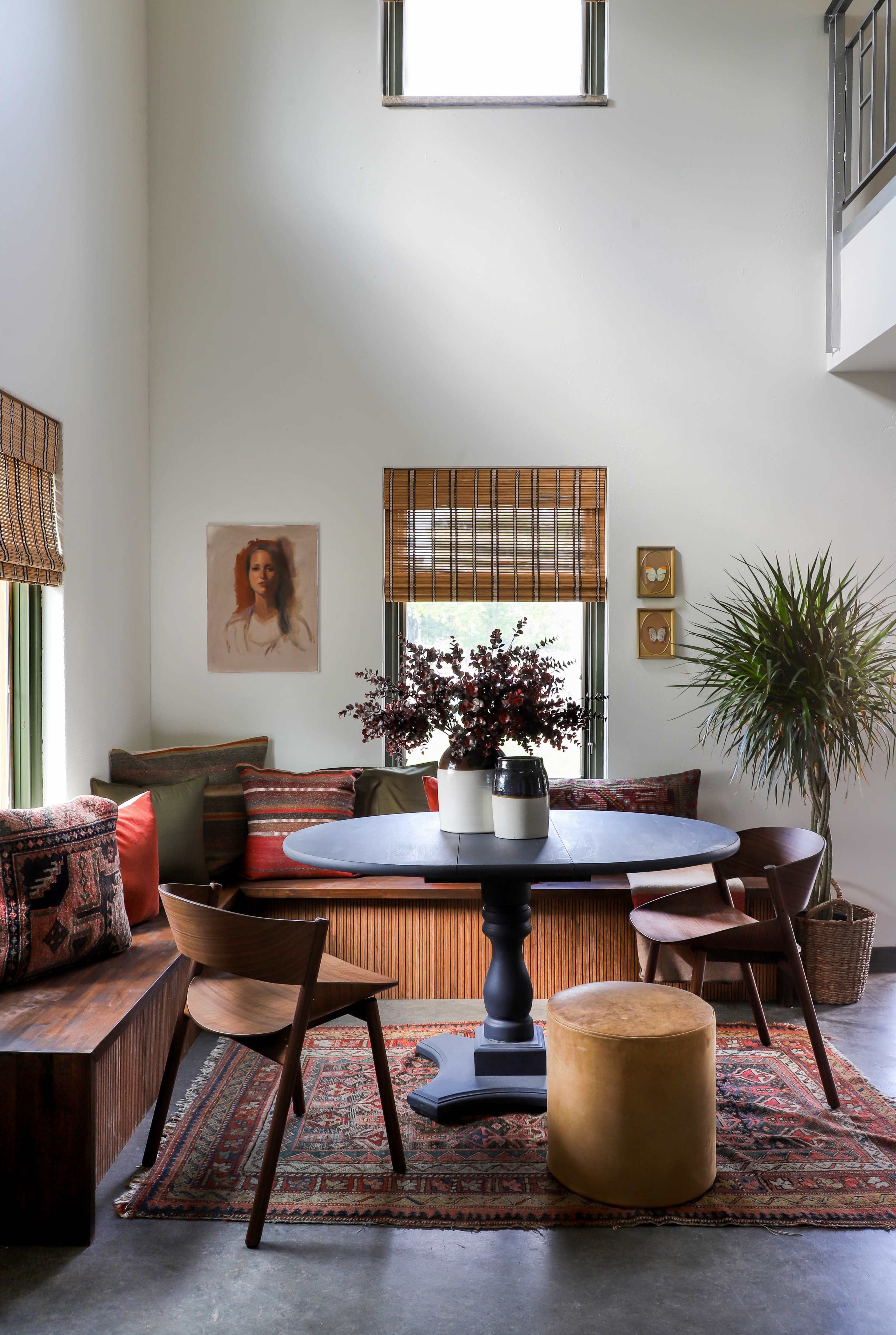 A cozy dining area features a round black table surrounded by wooden chairs and a cushioned bench, adorned with decorative pillows and plants.